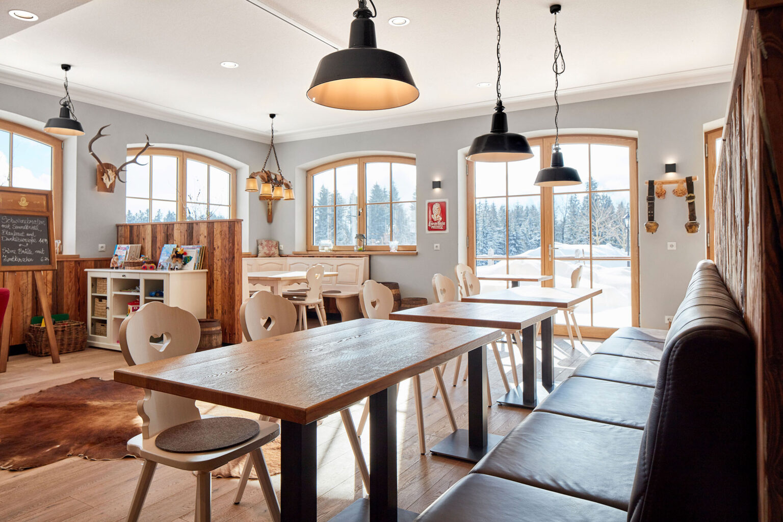 Ein gemütlich eingerichtetes Restaurant mit großen Fenstern, durch die eine schneebedeckte Landschaft zu sehen ist. Der Raum ist mit Holztischen und weißen Stühlen mit herzförmigen Rückenlehnen ausgestattet. An der Wand hängen rustikale Dekorationen, wie ein Hirschgeweih und traditionelle Kuhglocken. Schwarze Hängelampen sorgen für eine warme Beleuchtung. Ein Regal mit Büchern und ein Schild mit Menüangeboten befinden sich ebenfalls im Raum.