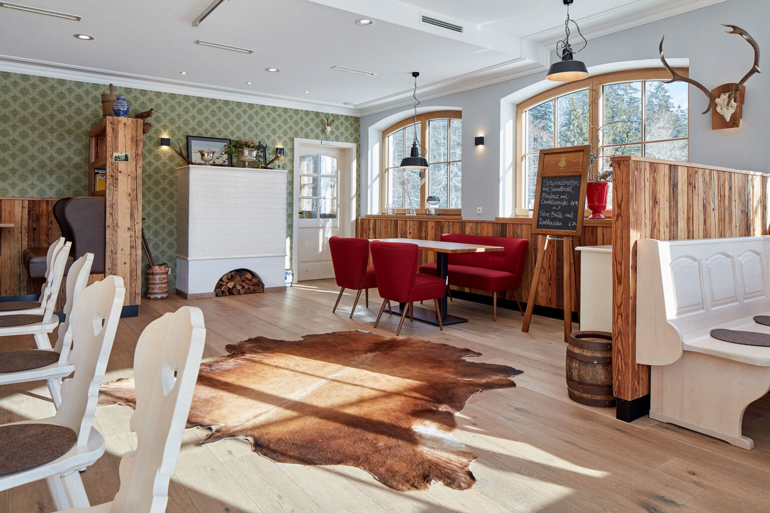 Ein gemütliches, traditionell eingerichtetes Restaurant mit Holzböden und Holzmöbeln. An den Wänden hängen Jagdtrophäen und Dekorationen. Es gibt weiße Stühle, rote Polstersessel an einem Holztisch und eine weiße Ofenheizung mit Holzvorrat darunter. Ein großes Kuhfell liegt auf dem Boden. Große Fenster lassen viel Tageslicht in den Raum. In der Ecke steht eine Tafel mit handgeschriebenen Menüspezialitäten.