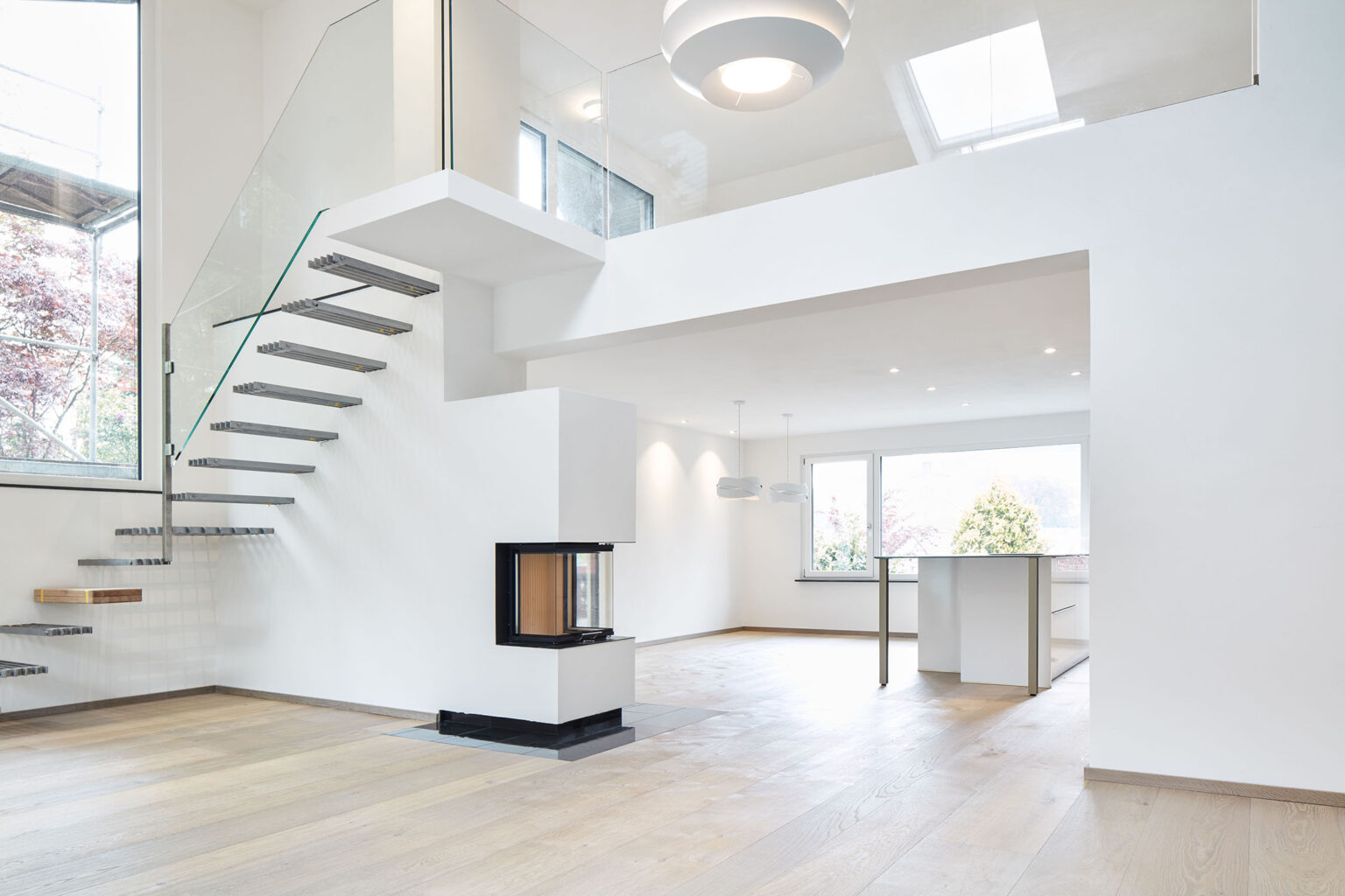 Ein modernes, minimalistisches Wohninterieur mit einer offenen Raumgestaltung. Eine freischwebende Treppe mit Metallstufen und einem Glasgeländer führt in das Obergeschoss. Unter der Treppe befindet sich ein moderner Kamin mit Glasfront. Der Boden ist aus hellem Holz, und die Wände sind weiß gestrichen. Große Fenster lassen viel natürliches Licht in den Raum. Im Hintergrund befindet sich ein Essbereich mit einem hohen Tisch und hängenden Lampen. Die Decke ist hoch und trägt zu einem großzügigen Raumgefühl bei.