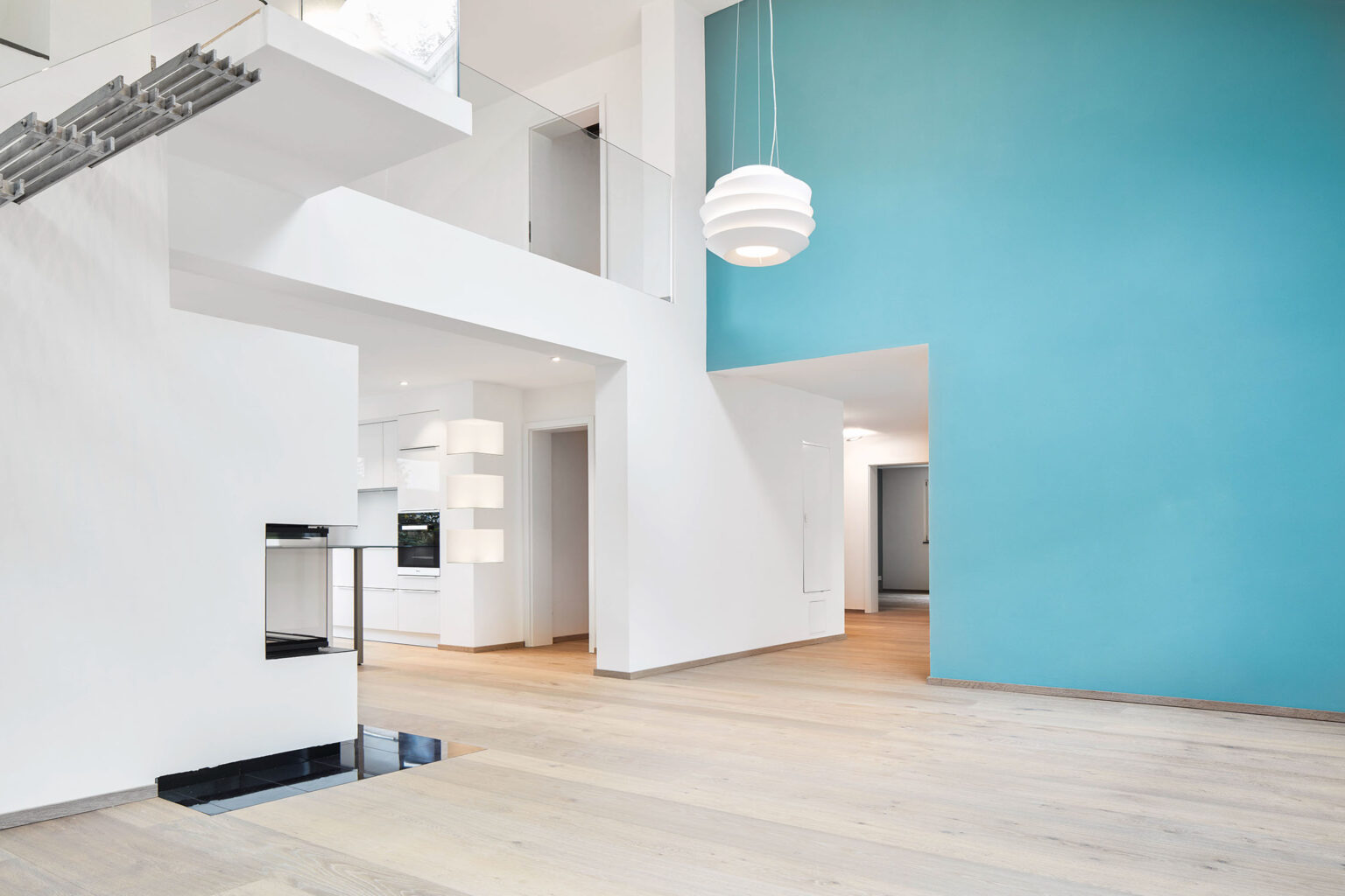 Ein modernes, minimalistisches Wohninterieur mit einer offenen Raumgestaltung. Eine freischwebende Treppe mit Metallstufen und einem Glasgeländer führt in das Obergeschoss. Unter der Treppe befindet sich ein moderner Kamin mit Glasfront. Der Boden ist aus hellem Holz, und die Wände sind weiß gestrichen, wobei eine Wand in einem auffälligen Blau gehalten ist. Große Fenster lassen viel natürliches Licht in den Raum. Im Hintergrund ist eine offene Küche mit weißen Schränken und modernen Geräten zu sehen. Eine stilvolle Hängelampe beleuchtet den Raum.