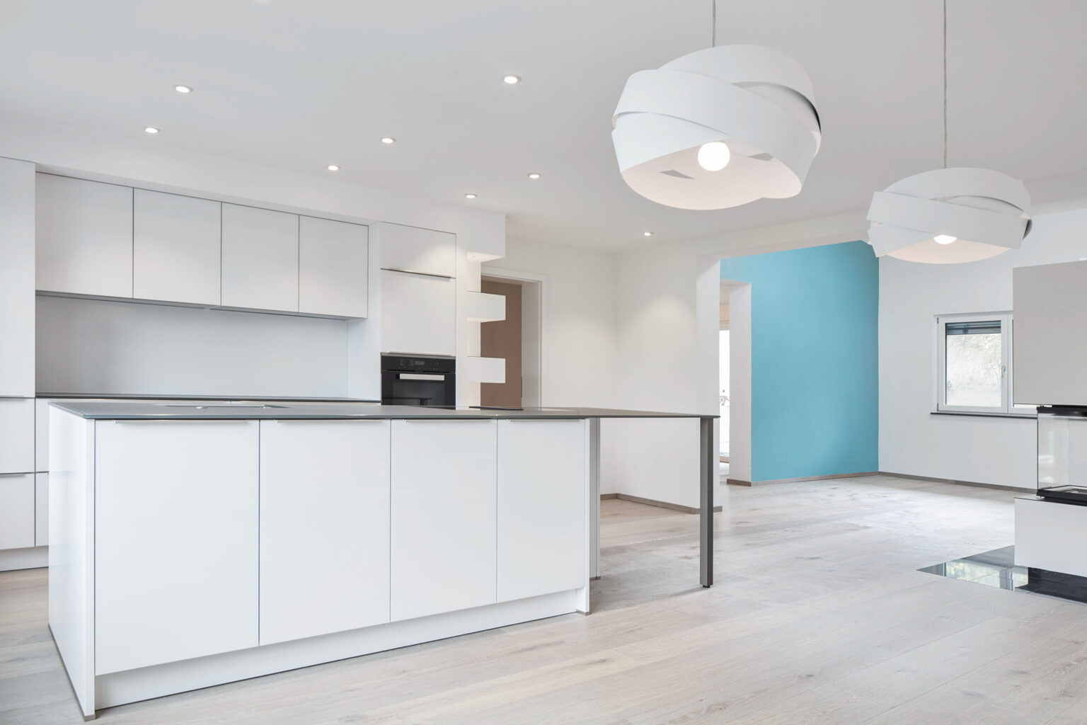 Eine moderne, minimalistische Küche mit weißen Schränken und einer großen Kücheninsel. Die Küche ist in einem offenen Raum gestaltet, der von zwei modernen weißen Hängelampen beleuchtet wird. Die Wände sind weiß gestrichen, mit einer auffälligen blauen Akzentwand im Hintergrund. Der Boden ist aus hellem Holz. Links im Bild befindet sich eine Tür, die in einen anderen Raum führt, und rechts ist ein Fenster zu sehen, das zusätzliches natürliches Licht hereinlässt. Die Deckenbeleuchtung besteht aus eingebauten Strahlern, die den Raum gut ausleuchten.
