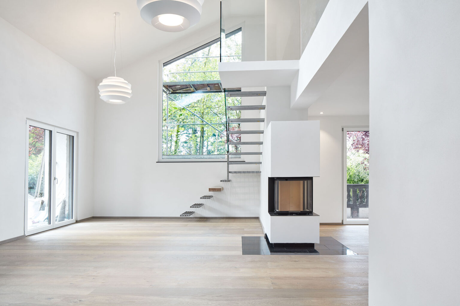 Ein modernes, minimalistisches Wohninterieur mit einer offenen Raumgestaltung und hohen Decken. Eine freischwebende Treppe mit Metallstufen und einem Glasgeländer führt in das Obergeschoss. Unter der Treppe befindet sich ein moderner Kamin mit Glasfront. Der Boden ist aus hellem Holz, und die Wände sind weiß gestrichen. Große Fenster und eine Glastür lassen viel natürliches Licht in den Raum und bieten einen Blick ins Freie. Zwei moderne, weiße Hängelampen hängen von der Decke und beleuchten den Raum.