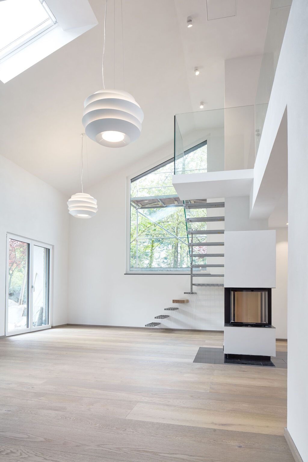 Ein modernes, minimalistisches Wohninterieur mit einer offenen Raumgestaltung und hohen Decken. Eine freischwebende Treppe mit Metallstufen und einem Glasgeländer führt in das Obergeschoss. Unter der Treppe befindet sich ein moderner Kamin mit Glasfront. Der Boden ist aus hellem Holz, und die Wände sind weiß gestrichen. Große Fenster und eine Glastür lassen viel natürliches Licht in den Raum und bieten einen Blick ins Freie. Zwei moderne, weiße Hängelampen beleuchten den Raum von der hohen Decke herab