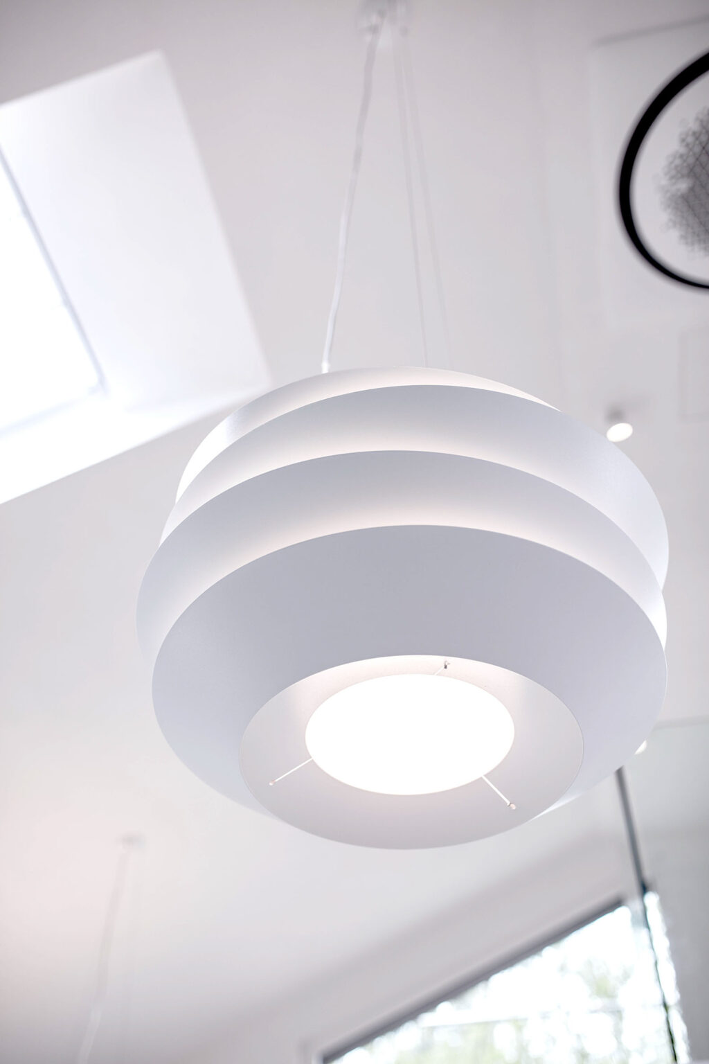 Eine Nahaufnahme einer modernen, weißen Hängelampe mit einem einzigartigen, geschichteten Design. Die Lampe hängt von der Decke eines hellen Raumes und strahlt weiches, gleichmäßiges Licht aus. Im Hintergrund sind die Umrisse eines Dachfensters und Teile des Raumes schemenhaft zu erkennen