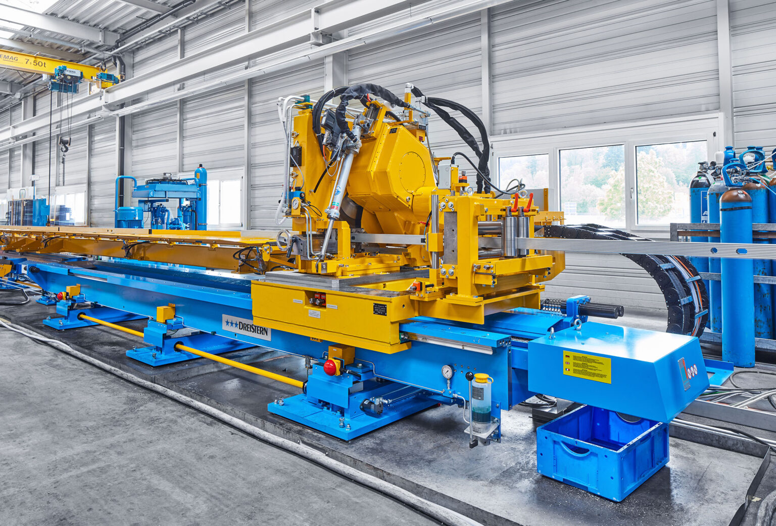 Eine große industrielle Maschine in einer Fabrikumgebung. Die Maschine hat einen gelben und blauen Rahmen und wird für die Bearbeitung von Metallstangen oder -profilen verwendet. Sie ist mit Hydraulikschläuchen, Steuerungen und mehreren beweglichen Teilen ausgestattet. Im Hintergrund sind weitere industrielle Geräte und Fenster zu sehen, die Tageslicht in die Halle lassen. Der Boden besteht aus Beton und die Umgebung wirkt sauber und gut organisiert.