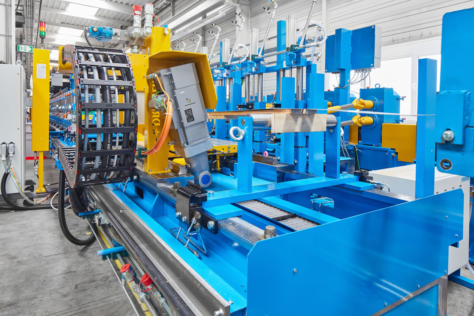 Eine große industrielle Maschine in einer Fabrikumgebung, die für die Bearbeitung oder den Transport von Metallprofilen verwendet wird. Die Maschine hat einen blauen Rahmen mit gelben Komponenten und ist mit verschiedenen mechanischen und elektrischen Bauteilen ausgestattet. Ein beweglicher Arm und eine Führungsschiene sind sichtbar, ebenso wie zahlreiche Kabel und Schläuche. Im Hintergrund sind weitere industrielle Geräte und Werkzeuge zu sehen. Die Umgebung ist sauber und gut organisiert, mit Betonboden und hellen Wänden.