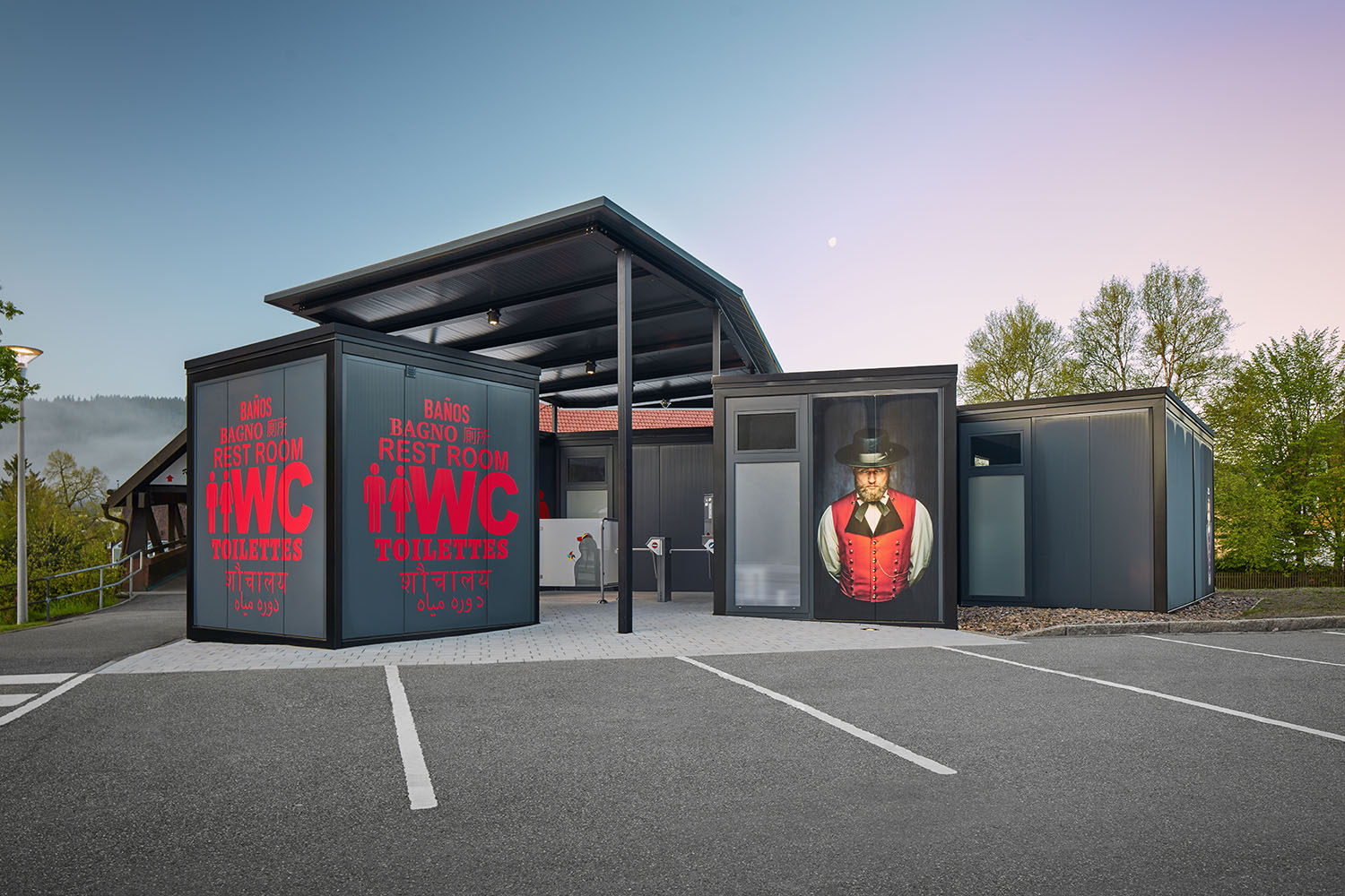 Ein modernes Toilettengebäude mit großen Schildern, auf denen in mehreren Sprachen 'WC', 'Toilettes', 'Rest Room' und 'Baños' steht. Auf der rechten Seite des Gebäudes ist ein großformatiges Bild eines Mannes in traditioneller Tracht zu sehen. Das Gebäude hat einen überdachten Eingangsbereich und steht auf einem gepflasterten Platz, umgeben von Bäumen und einem Parkplatz