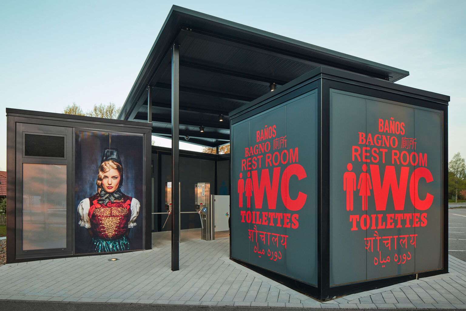 Ein modernes Toilettengebäude mit großen Schildern, auf denen in mehreren Sprachen 'WC', 'Toilettes', 'Rest Room' und 'Baños' steht. Auf der linken Seite des Gebäudes ist ein großformatiges Bild einer Frau in traditioneller Tracht zu sehen. Das Gebäude hat einen überdachten Eingangsbereich und steht auf einem gepflasterten Platz