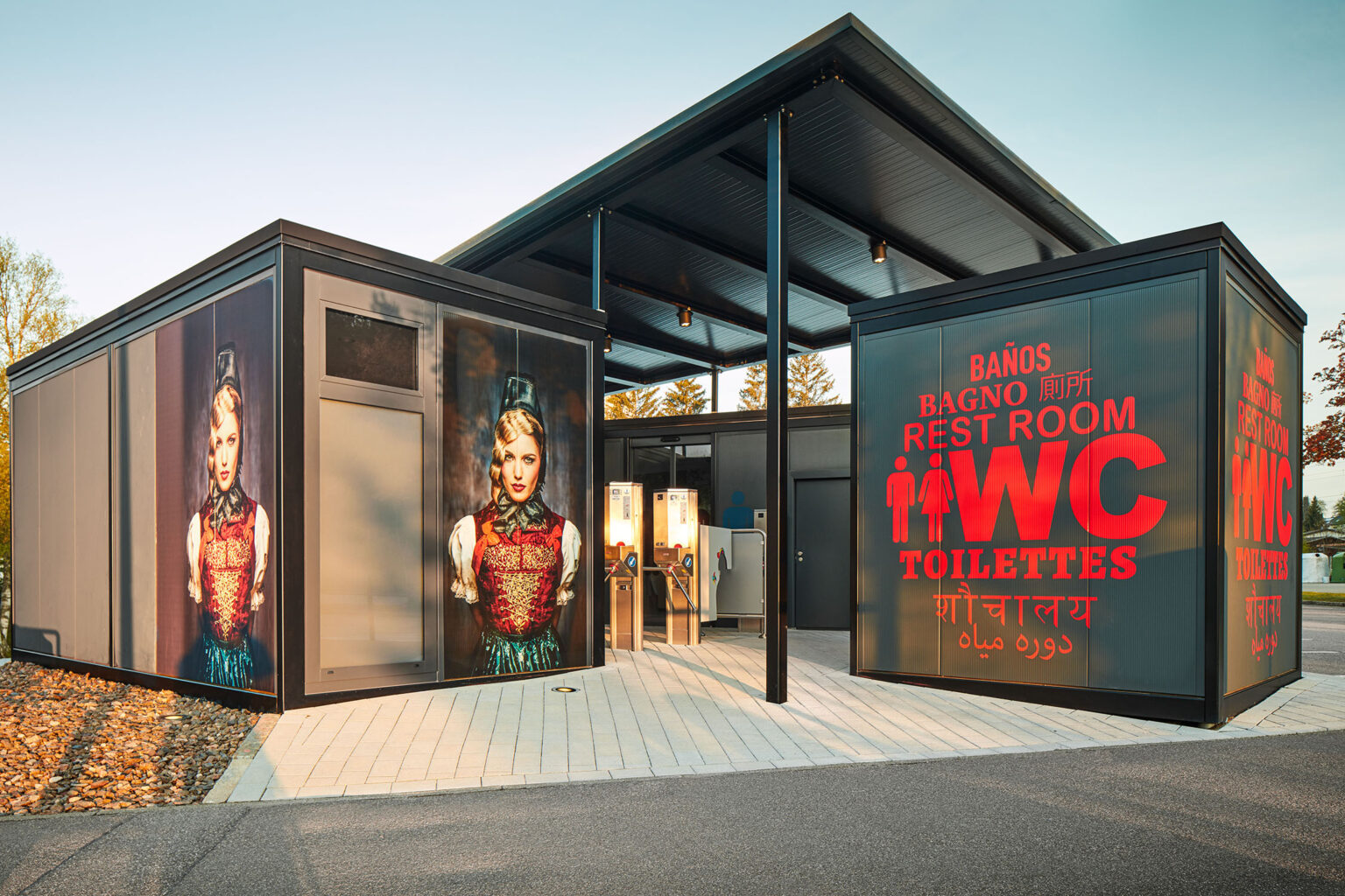 Ein modernes Toilettengebäude mit einem großen Schild, auf dem in mehreren Sprachen 'WC', 'Toilettes', 'Rest Room' und 'Baños' steht. Auf der linken Seite des Gebäudes sind großformatige Bilder einer Frau in traditioneller Tracht zu sehen. Das Gebäude hat einen überdachten Eingangsbereich und steht auf einem gepflasterten Platz, umgeben von Bäumen.