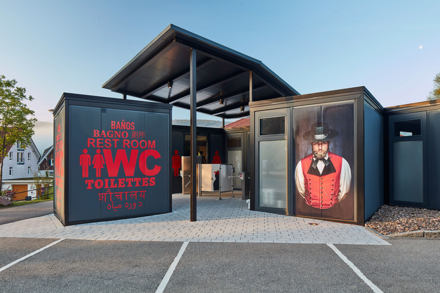 Ein modernes Toilettengebäude mit großen Schildern, auf denen in mehreren Sprachen 'WC', 'Toilettes', 'Rest Room' und 'Baños' steht. Auf der linken Seite des Gebäudes ist ein großformatiges Bild einer Frau in traditioneller Tracht zu sehen. Das Gebäude hat einen überdachten Eingangsbereich und steht auf einem gepflasterten Platz