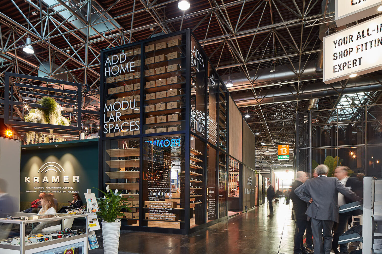Innenansicht einer modernen Messehalle mit hohen Decken und vielen Lichtern. Im Vordergrund sind Messestände zu sehen, darunter ein großer Stand von 'KRAMER', der für Ladenbau, Kühlraumbau und Modulbau wirbt. Mehrere Menschen sind in Gespräche vertieft und laufen durch die Halle. Der Stand hat ein Regal mit vielen kleinen Boxen und Pflanzen zur Dekoration.