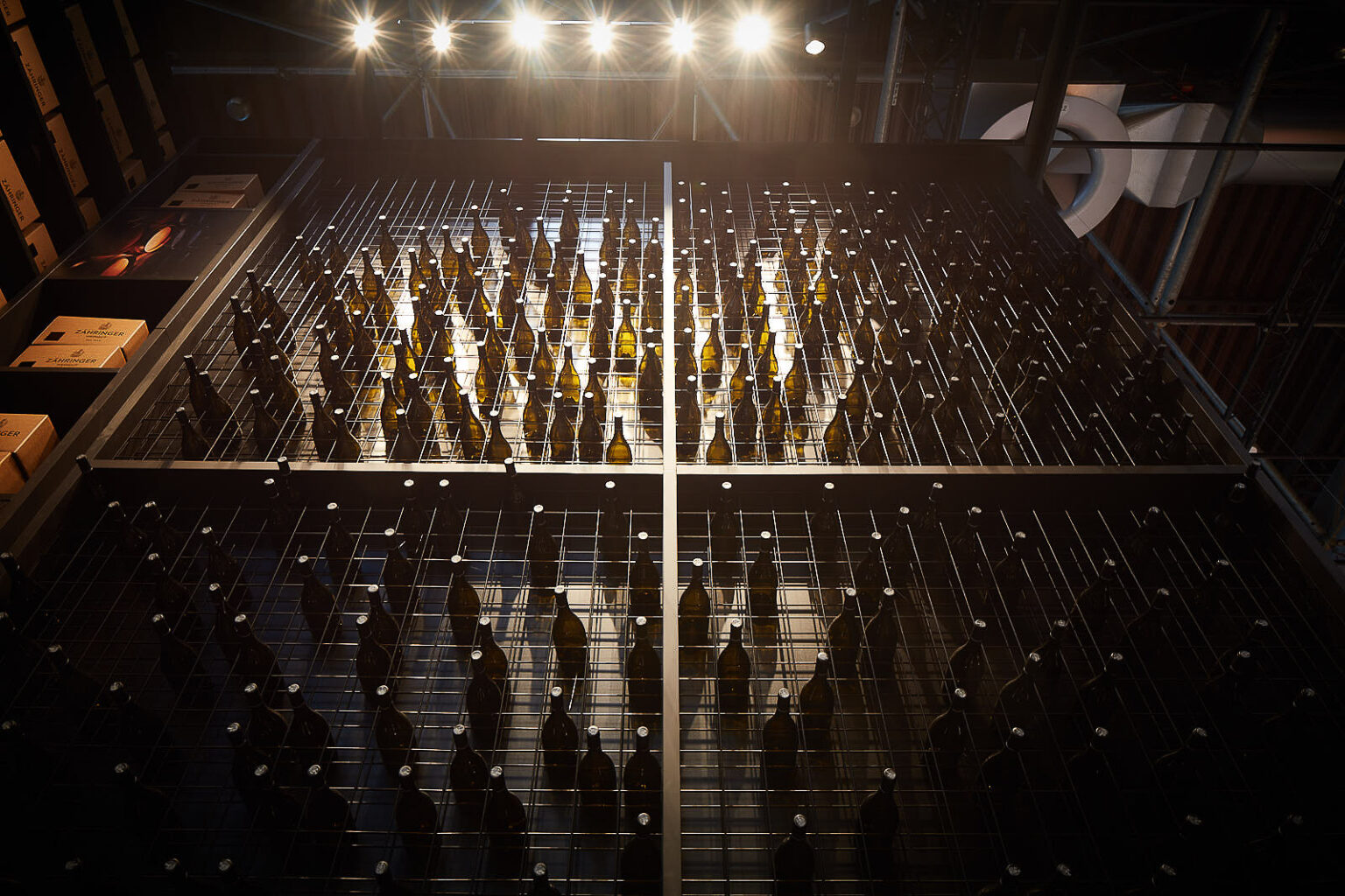 Blick von unten auf ein großes Regal mit zahlreichen Weinflaschen, die in einem Gittermuster angeordnet sind. Über dem Regal sind mehrere helle Strahler angebracht, die die Flaschen beleuchten. Links im Bild sind einige Kartons mit der Aufschrift 'ZÄHRINGER' zu sehen.