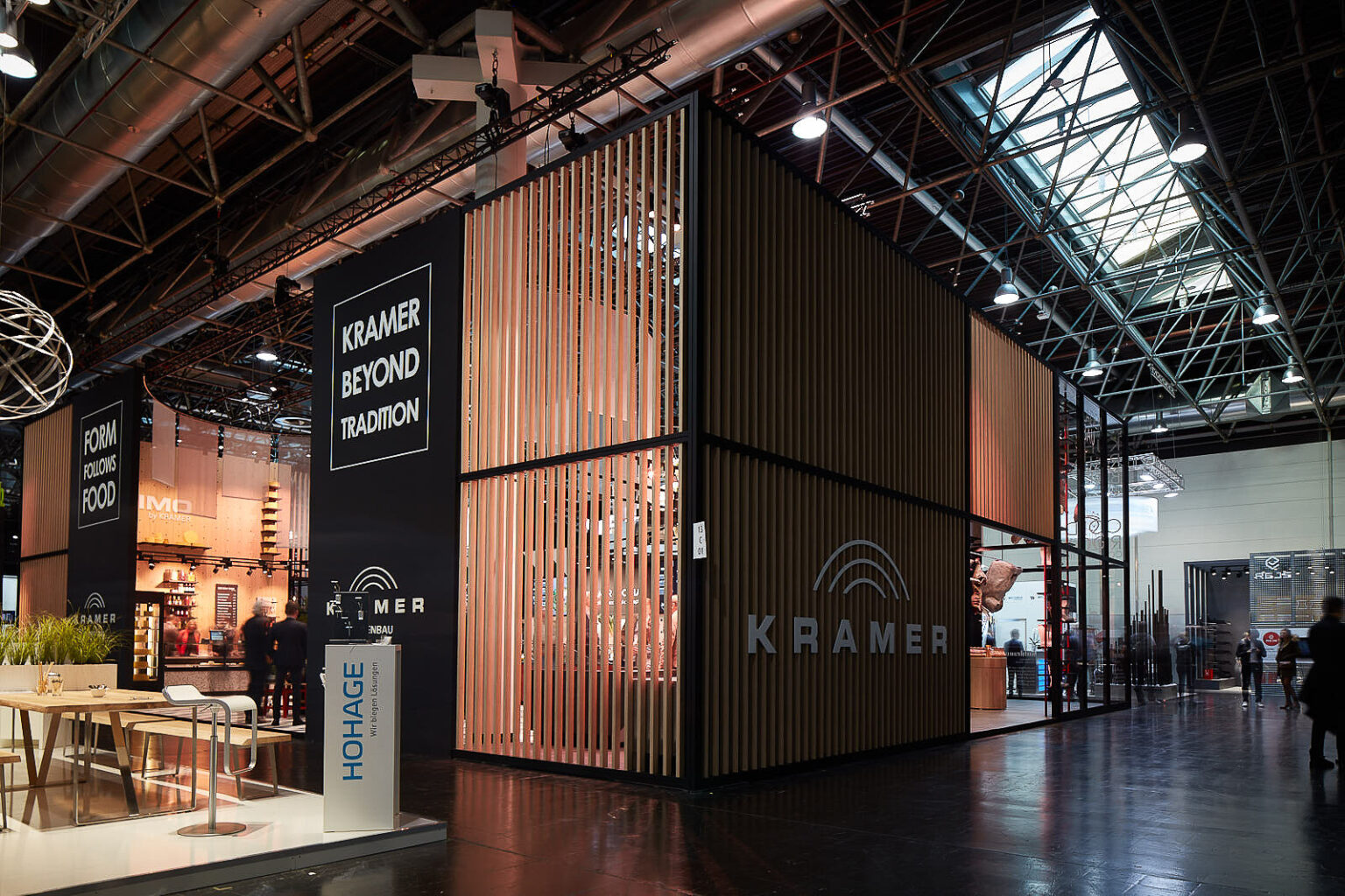 Ein großer Messestand von 'KRAMER' auf einer Messe. Der Stand ist modern gestaltet mit hohen, lamellenartigen Wänden und großen Schildern, auf denen 'KRAMER BEYOND TRADITION' und 'FORM FOLLOWS FOOD' steht. Im Vordergrund befindet sich ein Tisch mit Stühlen und ein Display mit der Aufschrift 'HOHAGE'. Der Stand ist gut beleuchtet und es sind mehrere Menschen zu sehen, die sich in der Halle bewegen.