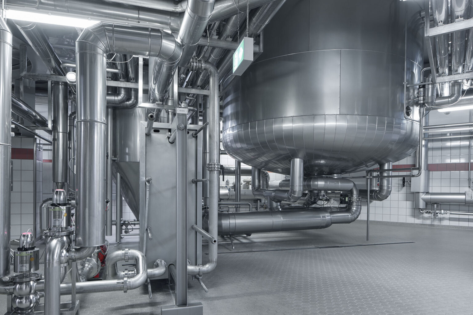 Ein industrieller Bereich mit einem großen, runden Edelstahlbehälter, der von einem komplexen Netzwerk aus Rohrleitungen umgeben ist. Die Rohrleitungen verlaufen in verschiedenen Richtungen und sind sauber installiert. Der Boden ist mit rutschfesten Platten ausgelegt und die Wände sind mit weißen Fliesen verkleidet. Ein Notausgangsschild ist an der Decke befestigt, was auf Sicherheitsvorkehrungen in der Anlage hinweist. Die gesamte Umgebung wirkt modern, sauber und gut organisiert.