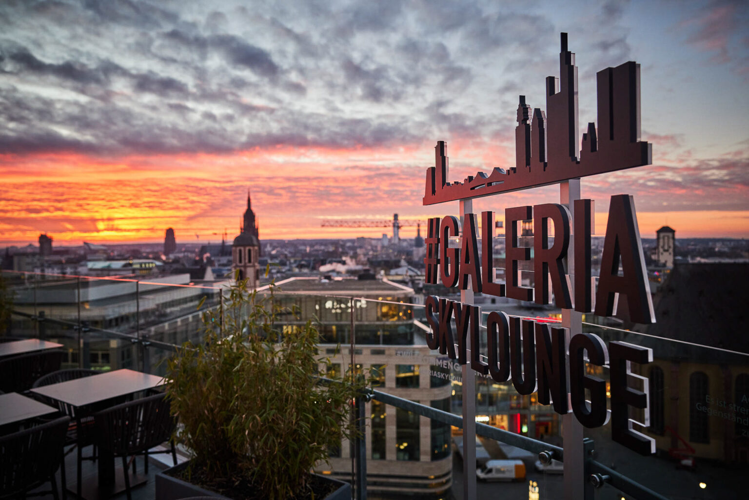 Blick auf eine Stadtlandschaft bei Sonnenuntergang von einer Dachterrasse aus. Im Vordergrund sind Tische und Stühle sowie Pflanzen zu sehen. Rechts im Bild steht ein Schild mit der Aufschrift '#GALERIA SKyLOUNGE'. Im Hintergrund sind Gebäude und ein farbenfroher Himmel mit Wolken zu sehen