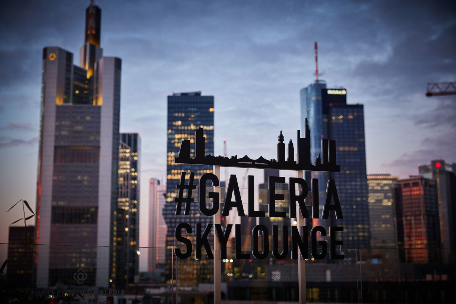 Blick auf die Skyline einer Stadt bei Dämmerung von einer Dachterrasse aus. Im Vordergrund ist ein Schild mit der Aufschrift '#GALERIA SKyLOUNGE' zu sehen. Im Hintergrund sind mehrere beleuchtete Hochhäuser vor einem dunkelblauen Himmel zu sehen.