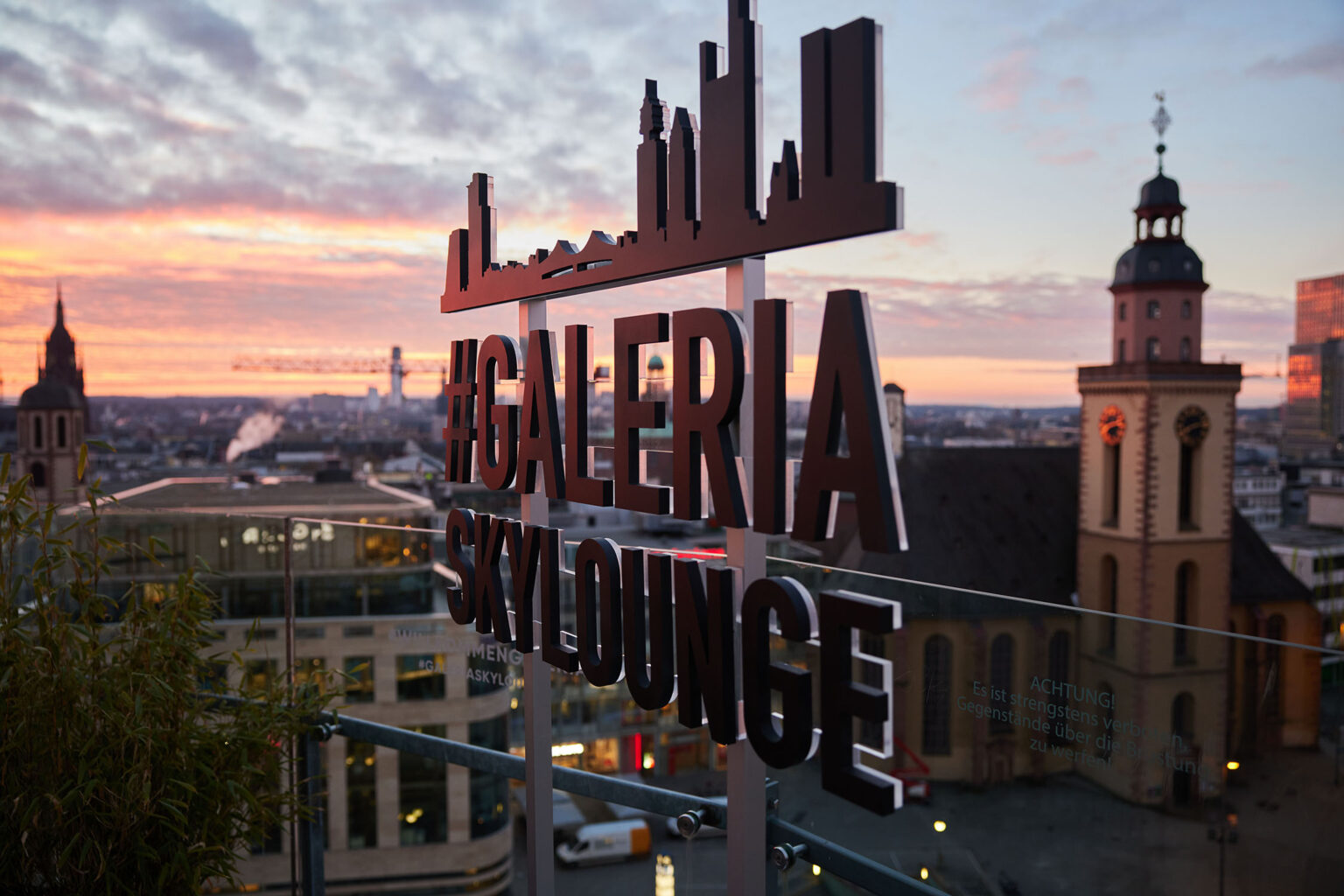 Blick auf eine Stadtlandschaft bei Sonnenuntergang von einer Dachterrasse aus. Im Vordergrund ist ein Schild mit der Aufschrift '#GALERIA SKyLOUNGE' zu sehen. Im Hintergrund sind mehrere Gebäude, darunter zwei Kirchtürme, sowie ein farbenfroher Himmel mit Wolken zu sehen.