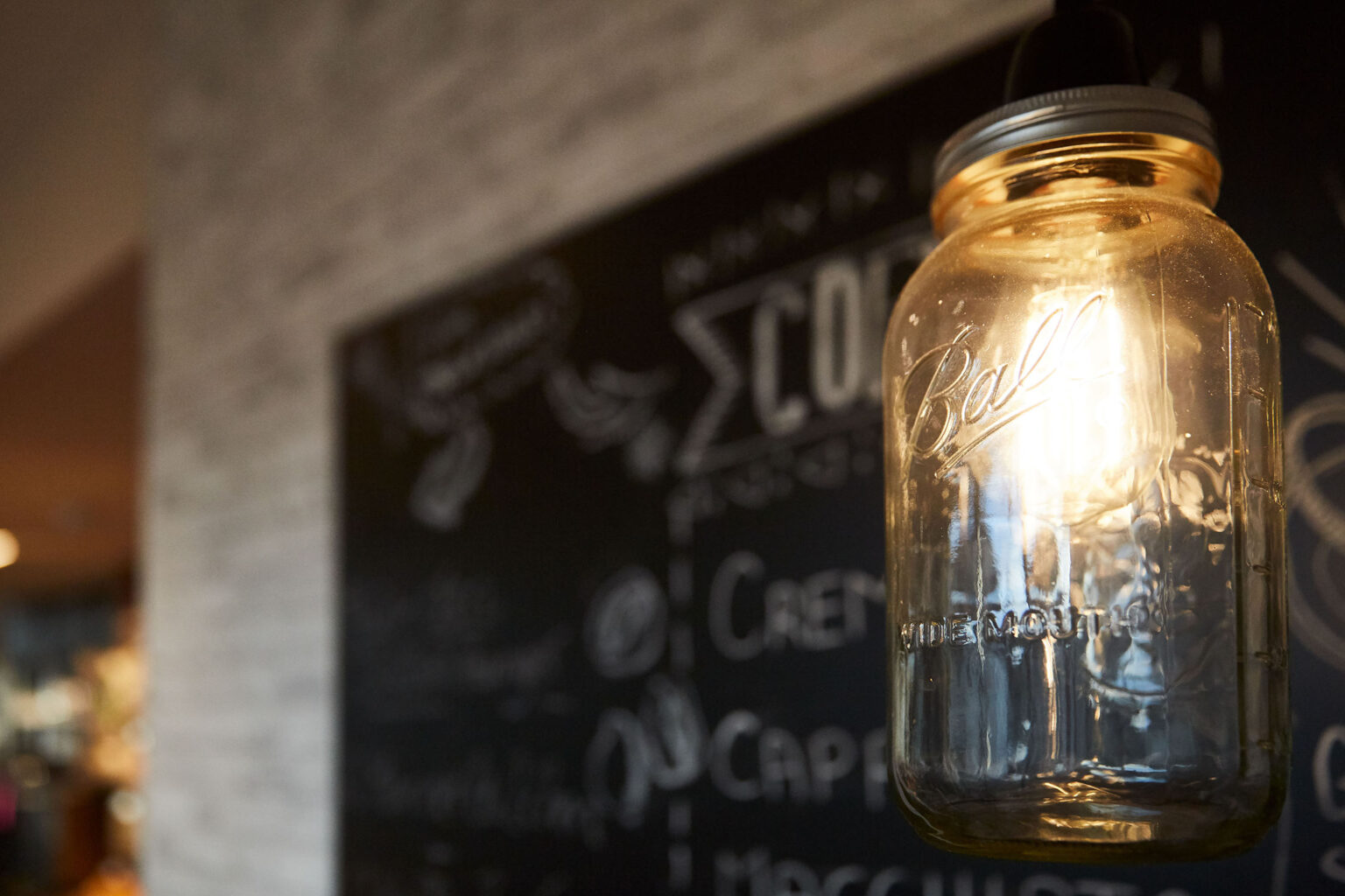 Eine Nahaufnahme einer hängenden Glühbirne, die in einem Einmachglas (Ball Wide Mouth) eingefasst ist. Im Hintergrund ist eine Wandtafel mit teilweise unscharfem Text und Zeichnungen zu sehen, die eine gemütliche, rustikale Atmosphäre in einem Café oder Restaurant vermittelt. Die Beleuchtung ist warm und schafft eine einladende Stimmung.