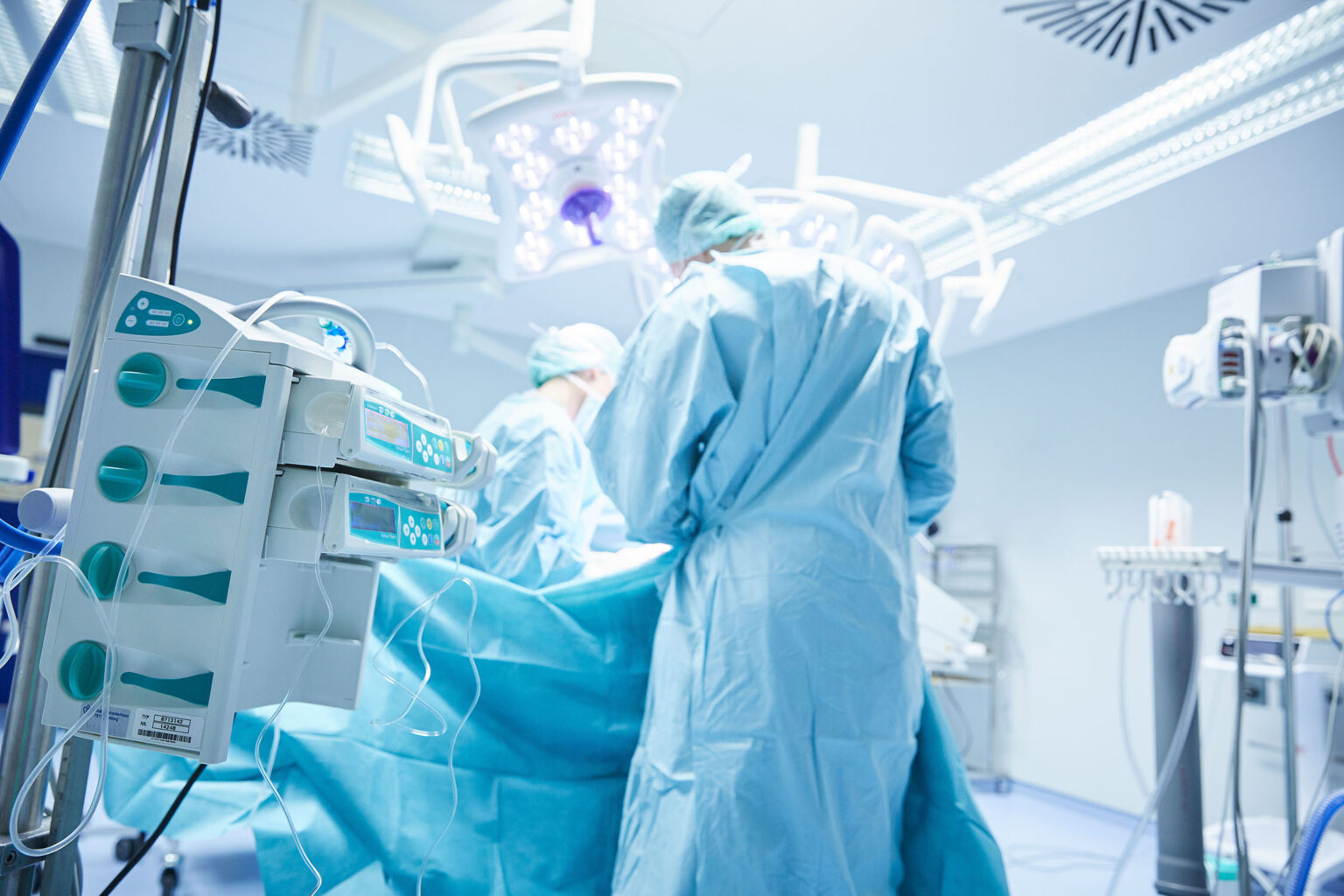 Ein Operationssaal mit medizinischem Personal und Ausrüstung. Im Vordergrund sind mehrere medizinische Geräte mit Schläuchen und Bildschirmen zu sehen. Im Hintergrund arbeiten Chirurgen in blauer OP-Kleidung und OP-Masken an einem Patienten unter hellem OP-Licht. Der Raum ist sauber und gut beleuchtet.