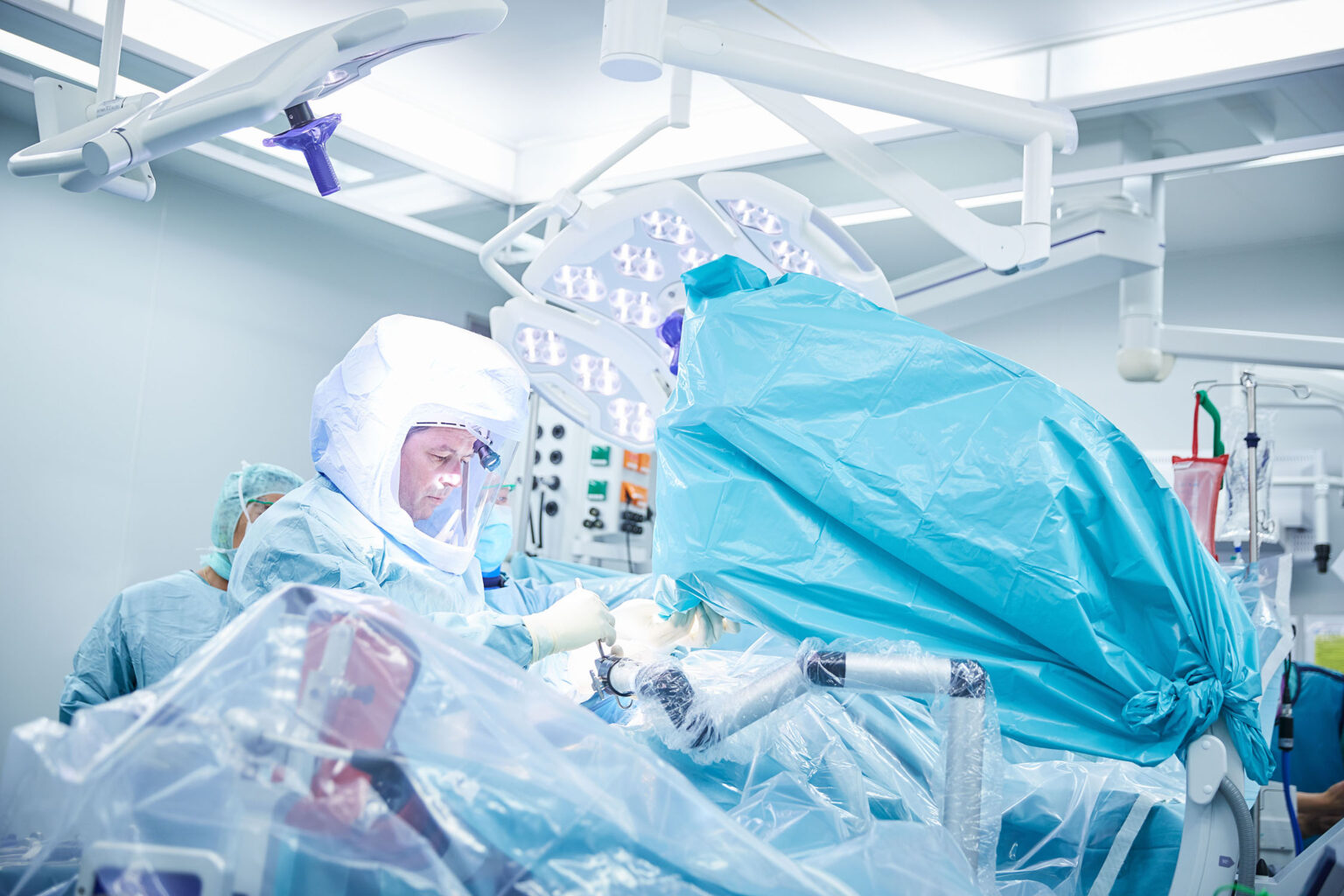 Ein Operationssaal mit einem Chirurgen, der eine spezielle Schutzhaube und OP-Kleidung trägt. Er arbeitet konzentriert an einem Patienten, der unter einem großen blauen Abdecktuch liegt. Weitere medizinische Geräte und Lampen sind im Hintergrund zu sehen, und das OP-Team ist in den Eingriff vertieft. Der Raum ist hell beleuchtet und mit moderner Ausrüstung ausgestattet