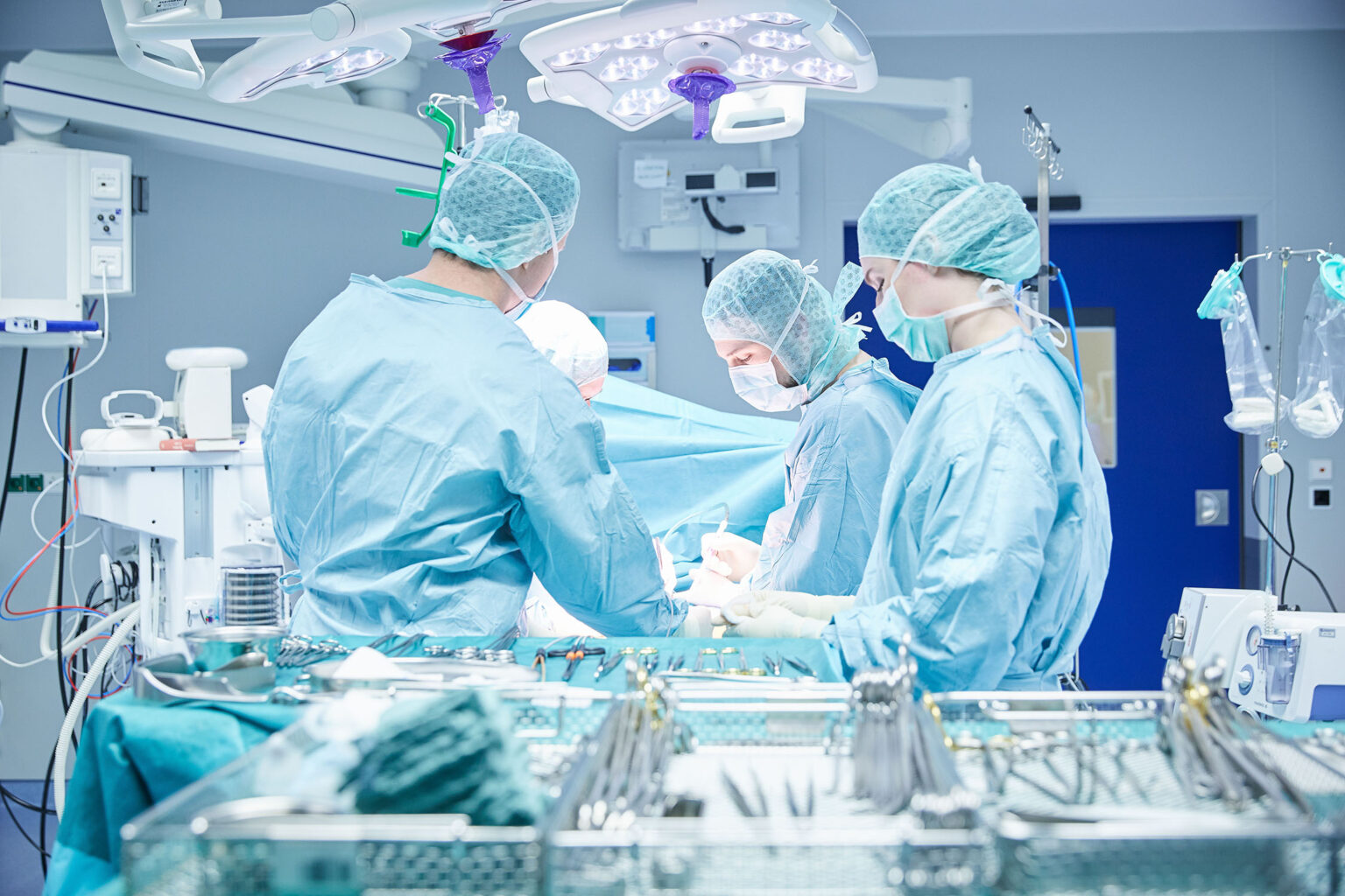 Ein Team von Chirurgen arbeitet in einem hell beleuchteten Operationssaal. Sie tragen blaue OP-Kleidung, OP-Masken und OP-Hauben. Im Vordergrund sind chirurgische Instrumente und Geräte zu sehen, die auf einem Tisch angeordnet sind. Die Chirurgen sind in den Eingriff vertieft und der Raum ist mit moderner medizinischer Ausrüstung ausgestattet. Helle OP-Leuchten hängen von der Decke.