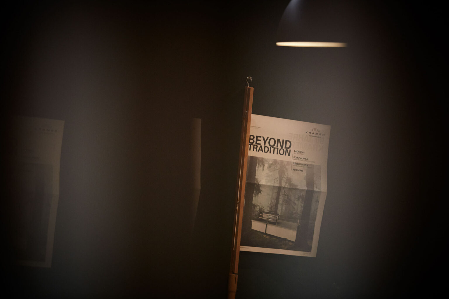 Ein Plakat mit der Aufschrift 'BEYOND TRADITION' hängt an einer dunklen Wand und wird von einem einzelnen Spotlicht beleuchtet. Auf dem Plakat ist auch der Name 'KRAMER' und eine Liste von Dienstleistungen wie Ladenbau, Kühlraumbau, Diamanttechnik und Addhome zu sehen. Im Hintergrund sind weitere unscharfe Plakate zu erkennen.