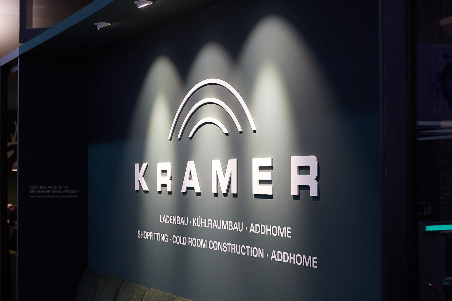 Eine beleuchtete Wand mit dem Logo und Schriftzug von 'KRAMER'. Unter dem Namen sind die Dienstleistungen aufgelistet: Ladenbau, Kühlraumbau, Addhome, Shopfitting, Cold Room Construction. Die Wand ist dunkel, und das Logo sowie die Schriftzüge sind in weiß gehalten.