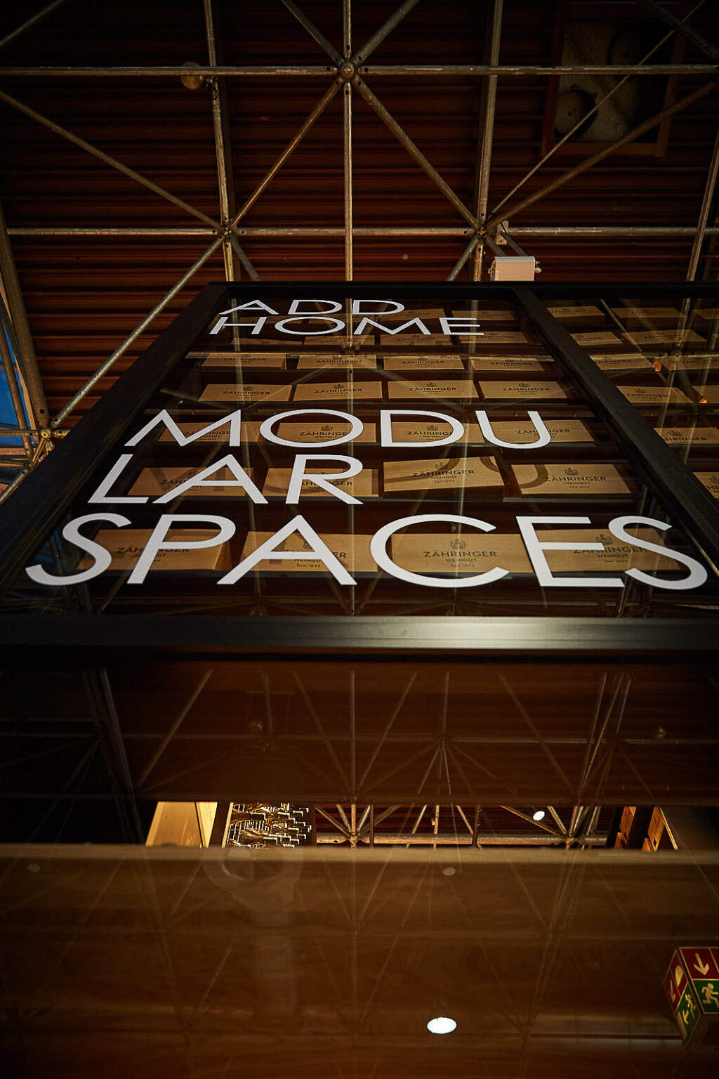 Blick von unten auf ein hohes Regal an einem Messestand mit der Aufschrift 'ADD HOME MODULAR SPACES'. Das Regal ist gefüllt mit vielen kleinen Boxen, die mit 'ZÄHRINGER' beschriftet sind. Über dem Regal ist das Dach der Messehalle mit Metallstreben und Beleuchtung zu sehen.