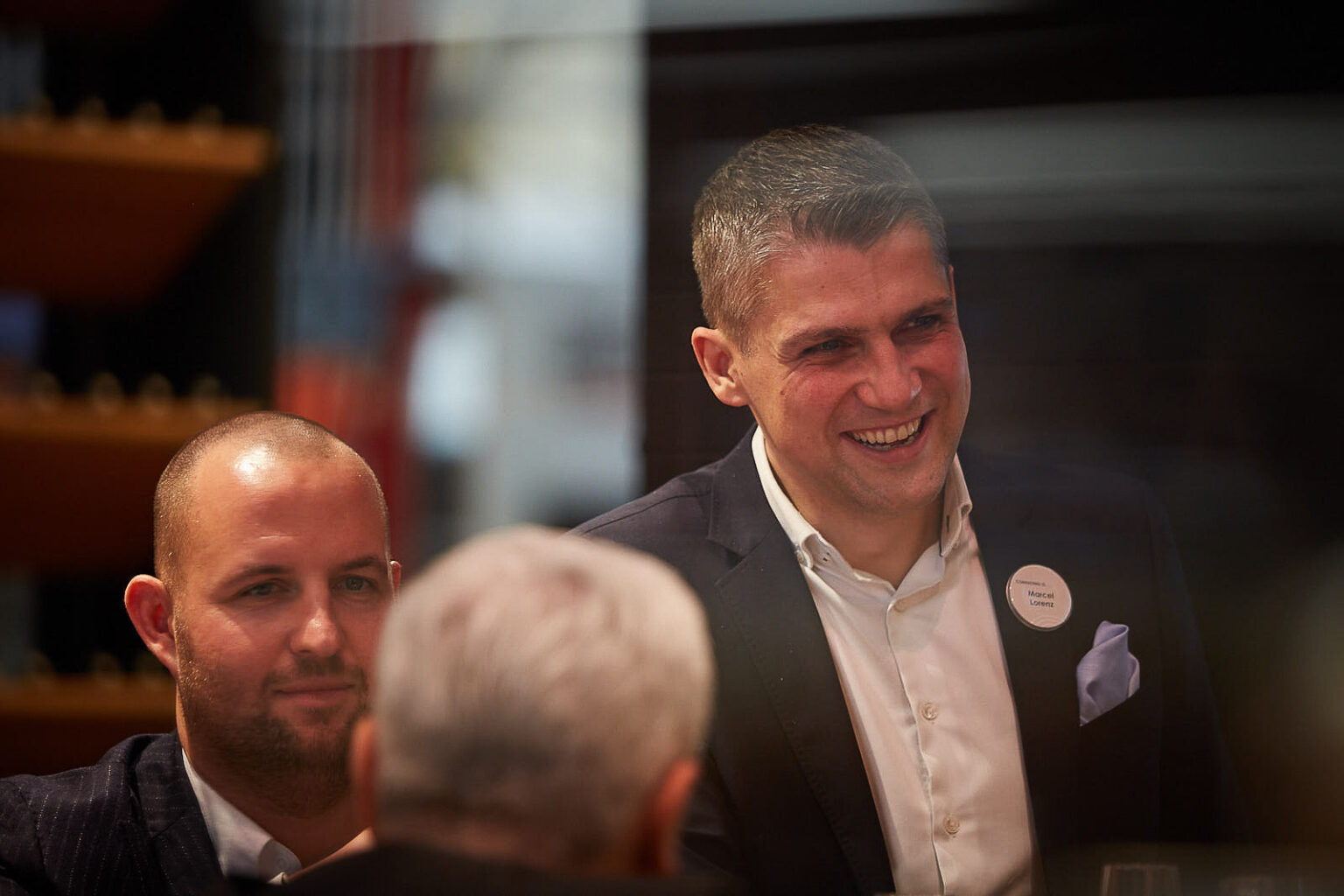 Zwei Männer in Businesskleidung sind in einem Gespräch vertieft. Der Mann rechts trägt ein dunkles Sakko und ein weißes Hemd und lächelt, er trägt ein Namensschild mit der Aufschrift 'Marcel Lorenz'. Der Mann links hat einen kurz geschnittenen Haarschnitt und trägt ebenfalls ein dunkles Sakko und ein weißes Hemd. Im Vordergrund ist der unscharfe Hinterkopf einer weiteren Person zu sehen. Die Szene findet in einem gut beleuchteten, modernen Innenraum statt.