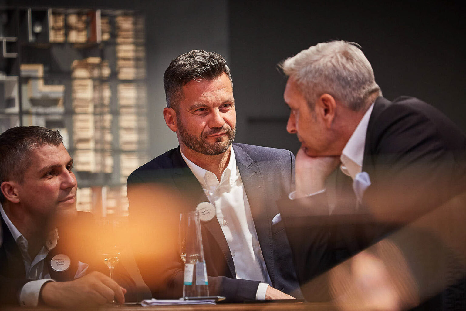 Drei Männer in Businesskleidung sitzen an einem Tisch und unterhalten sich. Der Mann in der Mitte hat dunkles, kurzes Haar und trägt ein dunkelblaues Sakko und ein weißes Hemd. Er trägt ein Namensschild mit der Aufschrift 'Thorsten ...'. Der Mann links trägt ein dunkles Sakko und ein weißes Hemd und hat ebenfalls ein Namensschild mit der Aufschrift 'Marcel Lorenz'. Der Mann rechts hat graues Haar und trägt ein dunkles Sakko und ein weißes Hemd. Die Szene spielt sich in einem gut beleuchteten Innenraum ab, im Hintergrund sind unscharfe Regale zu sehen.