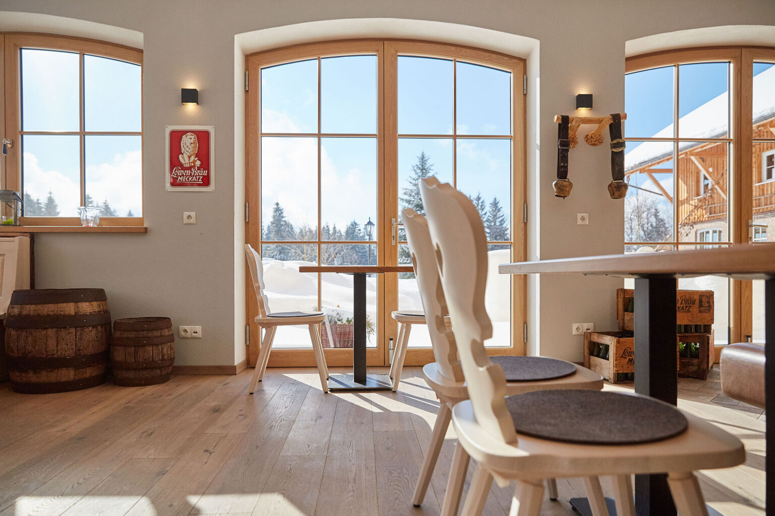 Ein heller, gemütlicher Raum mit großen Fenstern, die einen Blick auf eine schneebedeckte Landschaft bieten. Im Raum stehen weiße Holzstühle an Holztischen auf einem Holzboden. An der Wand hängt ein Löwenbräu Meckatz Schild, daneben sind zwei Holzfässer und an der Wand montierte Hirschgeweihe zu sehen. Der Raum wirkt einladend und traditionell.