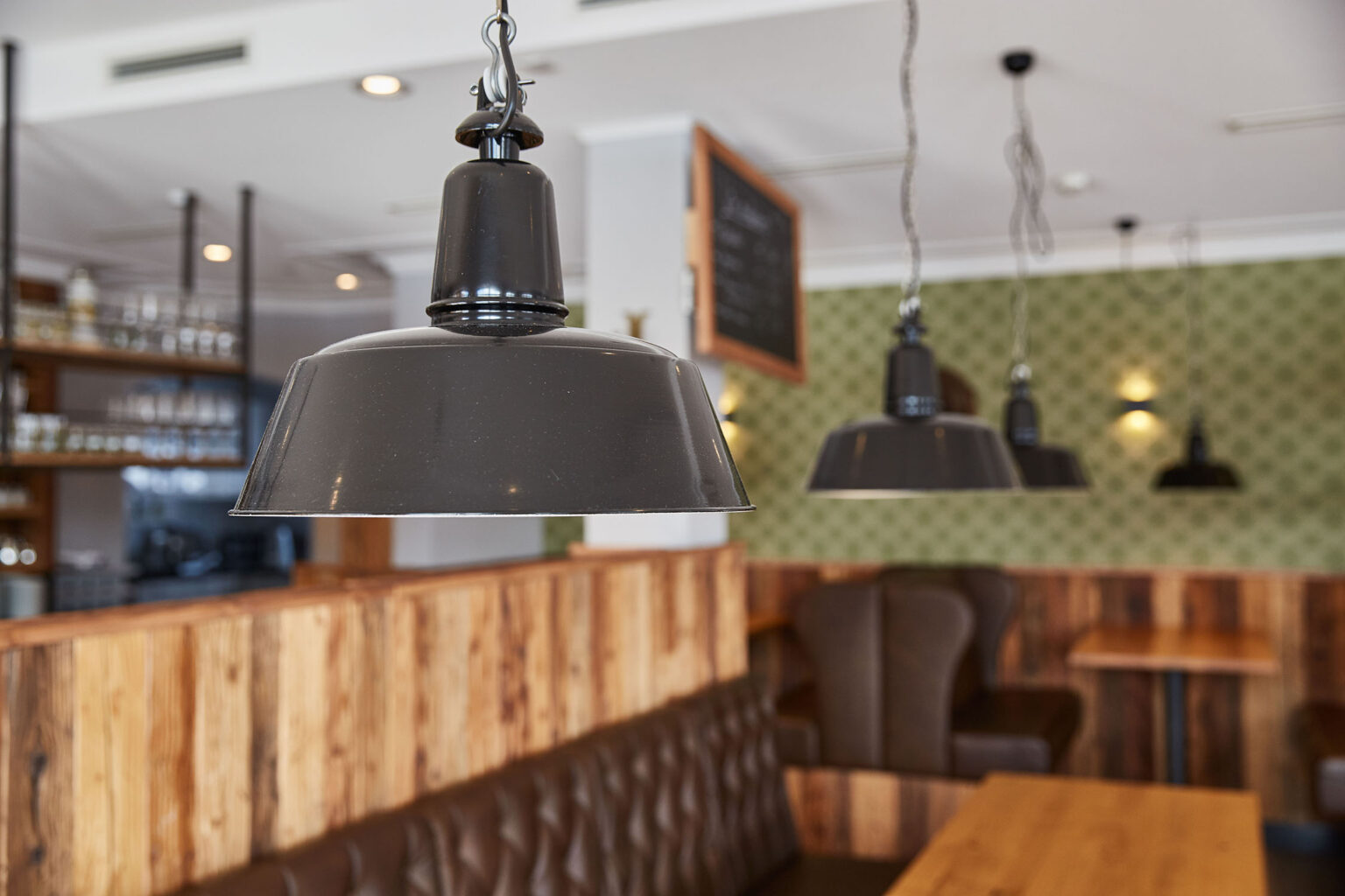 Ein stilvoll eingerichtetes Restaurant mit hängenden, schwarzen Metalllampen im Vordergrund. Im Hintergrund sind Holzbänke mit braunen Lederpolstern, Holztische und grün gemusterte Tapeten zu sehen. An der Wand hängt eine Tafel mit handgeschriebenen Menüangeboten. Der Raum wirkt gemütlich und einladend.