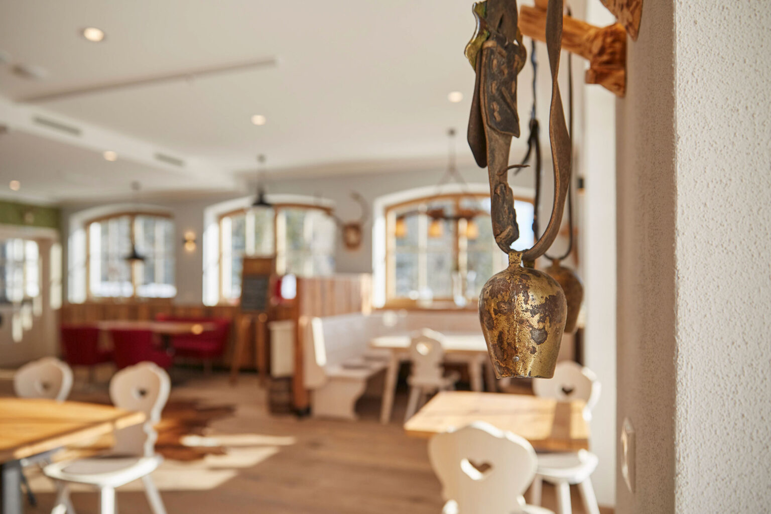 Ein helles, gemütliches Restaurant mit weißen Holzstühlen und Holztischen auf einem Holzboden. Im Vordergrund hängt eine rustikale Glocke an der Wand. Im Hintergrund sind große Fenster, durch die viel Tageslicht in den Raum fällt, sowie verschiedene Sitzbereiche mit roten und weißen Polsterbänken und Stühlen. Die Wände sind dekoriert mit traditionellen Elementen.