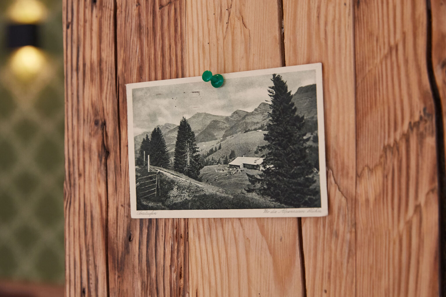 Eine schwarz-weiße Postkarte mit einem Landschaftsbild, das eine Bergszene mit Bäumen und einem Bauernhaus zeigt, ist mit einer grünen Pinnnadel an einer Holzwand befestigt. Im Hintergrund ist eine grün gemusterte Tapete sichtbar