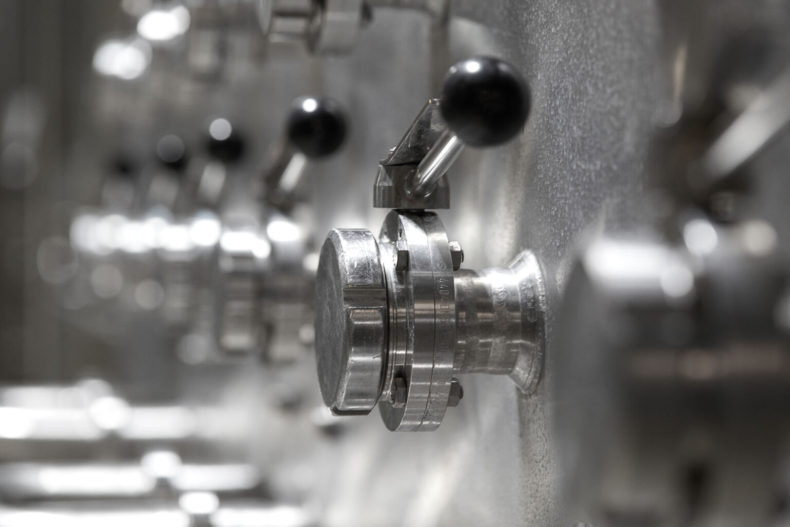 Eine Nahaufnahme von mehreren metallischen Ventilen und Hebeln an einer industriellen Maschine. Die Ventile sind präzise gefertigt und mit schwarzen Griffen ausgestattet. Die Umgebung ist unscharf, was den Fokus auf die Details der Ventile lenkt. Die reflektierenden Oberflächen der Metallteile vermitteln einen Eindruck von technischer Präzision und Sauberkeit.