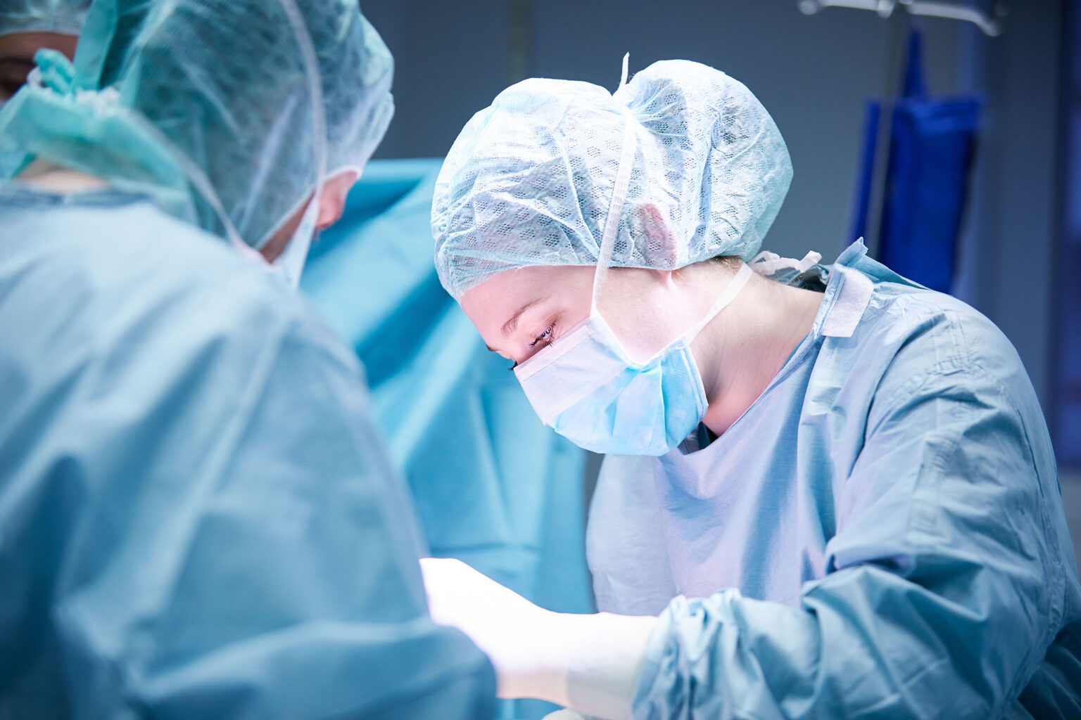 Ein Team von Chirurgen bei der Arbeit im Operationssaal. Im Vordergrund sind zwei Chirurgen zu sehen, die OP-Kleidung, OP-Masken und OP-Hauben tragen. Die Chirurgin auf der rechten Seite konzentriert sich auf die Operation, während sie nach unten blickt. Der Hintergrund zeigt weitere medizinische Ausrüstung und blaue Vorhänge. Die Szene ist hell beleuchtet.
