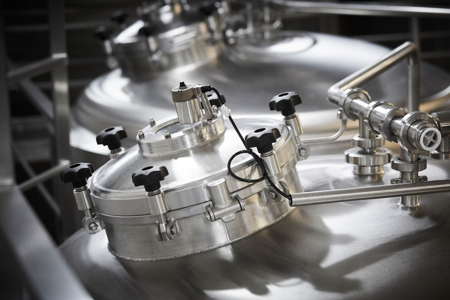 Eine Nahaufnahme von Edelstahlbehältern in einer industriellen Anlage, wahrscheinlich zur Verarbeitung von Flüssigkeiten oder Lebensmitteln. Die Behälter sind mit mehreren Ventilen, Verschlüssen und Rohrleitungen ausgestattet. Die Oberfläche der Behälter ist glänzend und reflektiert das Licht. Die Anordnung der Komponenten und die saubere Verarbeitung weisen auf eine moderne und hygienische Produktionsumgebung hin