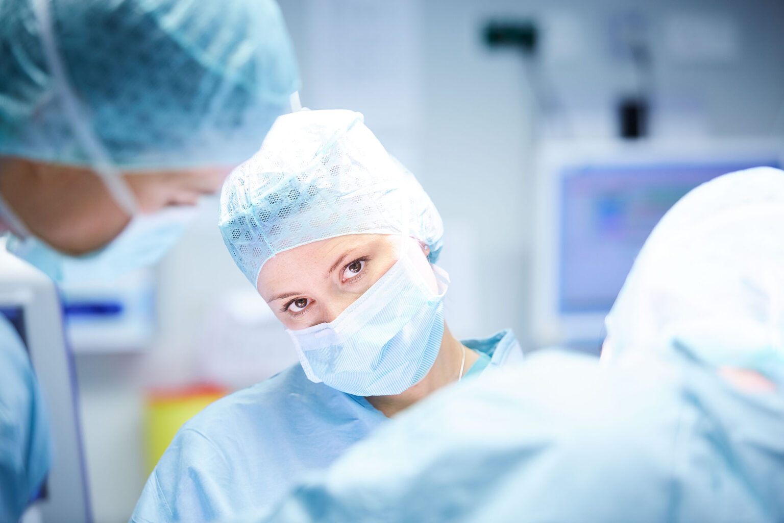 Ein Team von Chirurgen im Operationssaal. Im Vordergrund ist eine Chirurgin zu sehen, die eine blaue OP-Maske und eine blaue OP-Haube trägt. Sie schaut direkt in die Kamera. Weitere Chirurgen sind im Hintergrund unscharf zu sehen, während sie an einer Operation arbeiten. Der Raum ist hell beleuchtet und mit medizinischen Geräten ausgestattet.