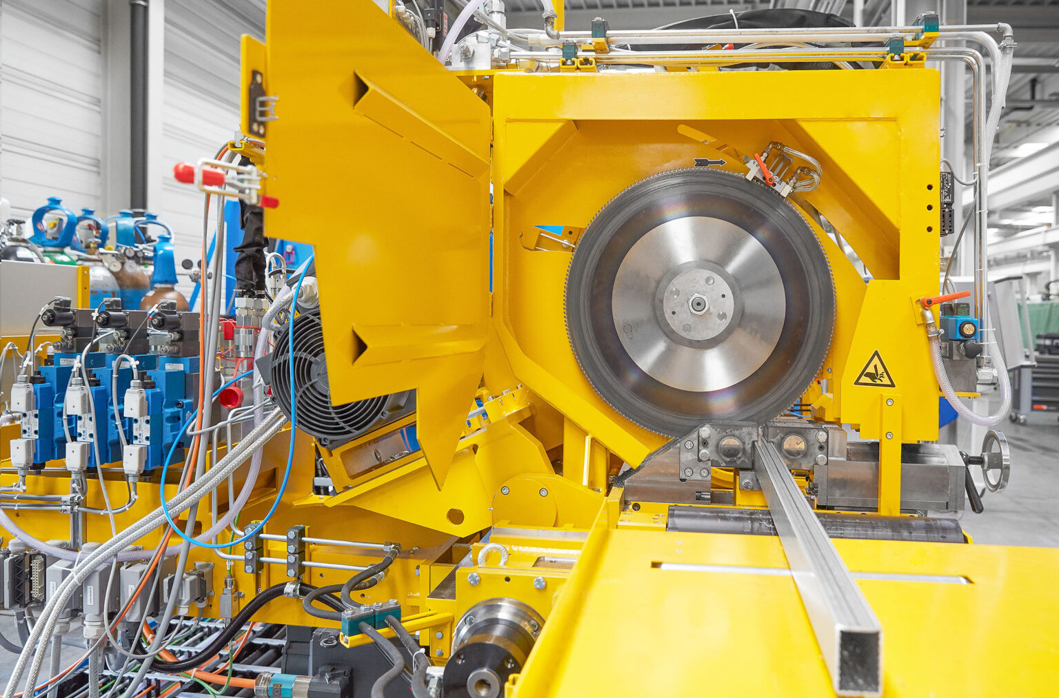 Eine Nahaufnahme einer gelben industriellen Maschine, die für das Schneiden von Metallstangen oder -profilen verwendet wird. Im Zentrum der Maschine befindet sich eine große Kreissäge, die von Hydraulik- und Elektrosystemen umgeben ist. Verschiedene Kabel und Schläuche sind an die Maschine angeschlossen. Im Hintergrund sind weitere Maschinen und industrielle Ausrüstung zu sehen. Die Umgebung wirkt sauber und gut organisiert.