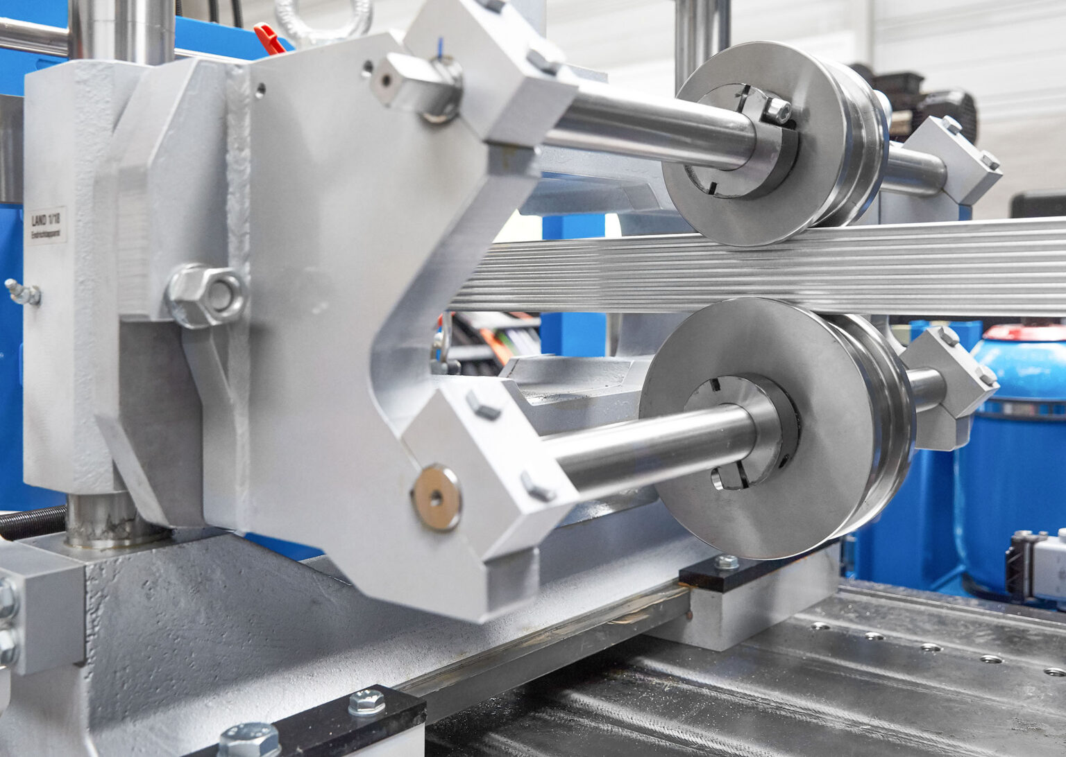 Eine Nahaufnahme einer industriellen Maschine mit mehreren Metallrollen und Stangen, die für die Bearbeitung oder den Transport von Materialien verwendet werden. Die Maschine besteht aus robustem Metall und ist präzise gefertigt. Im Hintergrund sind Teile der Fabrikumgebung und weitere Maschinen zu erkennen. Die gesamte Konstruktion vermittelt einen Eindruck von Stabilität und technischer Präzision.