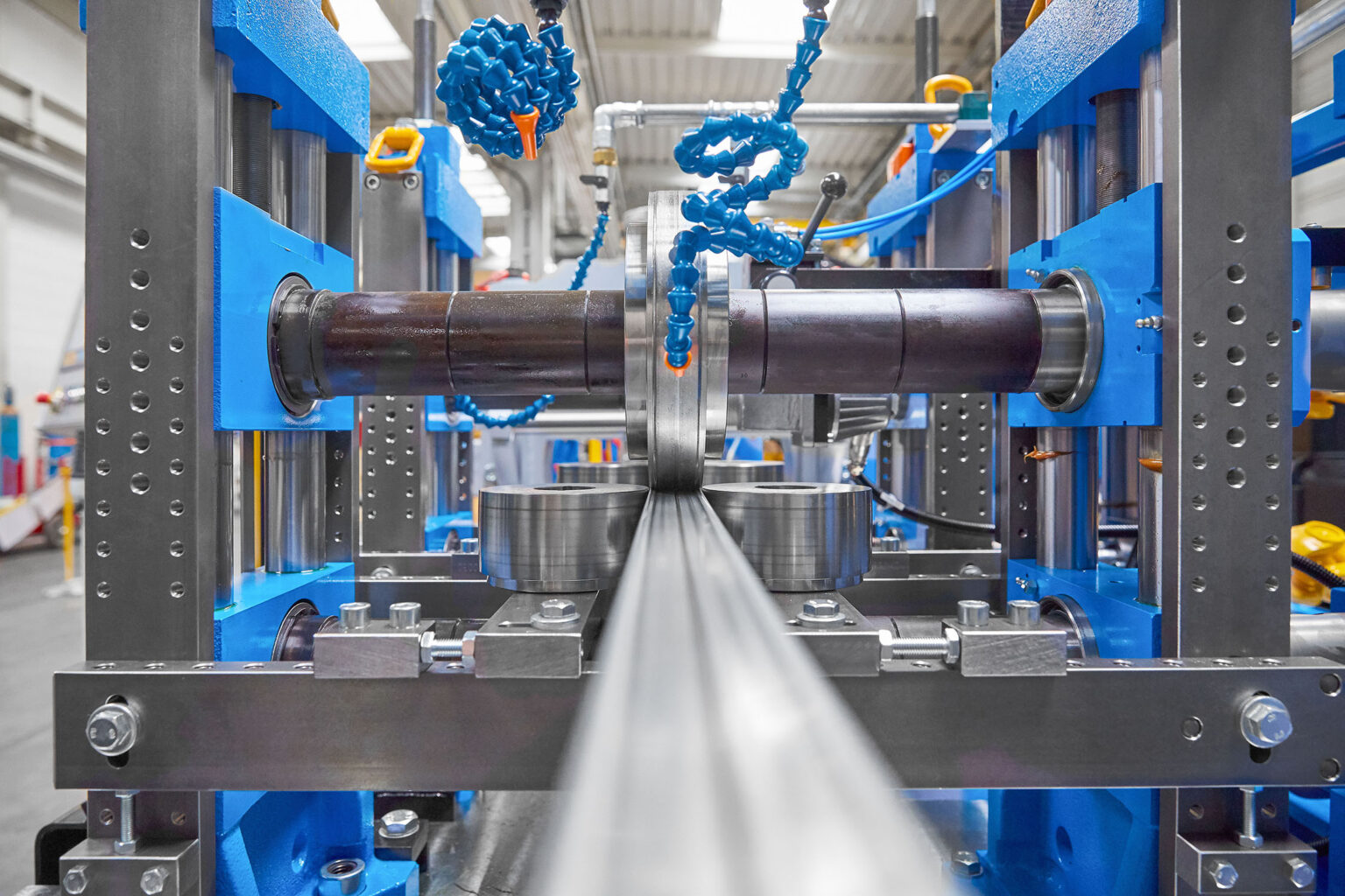 Eine Nahaufnahme einer industriellen Maschine, die für die Bearbeitung von Metallprofilen verwendet wird. Die Maschine hat einen blauen Rahmen und mehrere Rollen und Führungen, die das Metallprofil in Position halten. Flexible Kühlmitteldüsen sind sichtbar, die das Werkstück während des Bearbeitungsprozesses kühlen. Die Konstruktion ist robust und präzise, was auf eine hochwertige industrielle Fertigungsumgebung hinweist. Im Hintergrund sind weitere Maschinenteile und die industrielle Umgebung zu sehen.