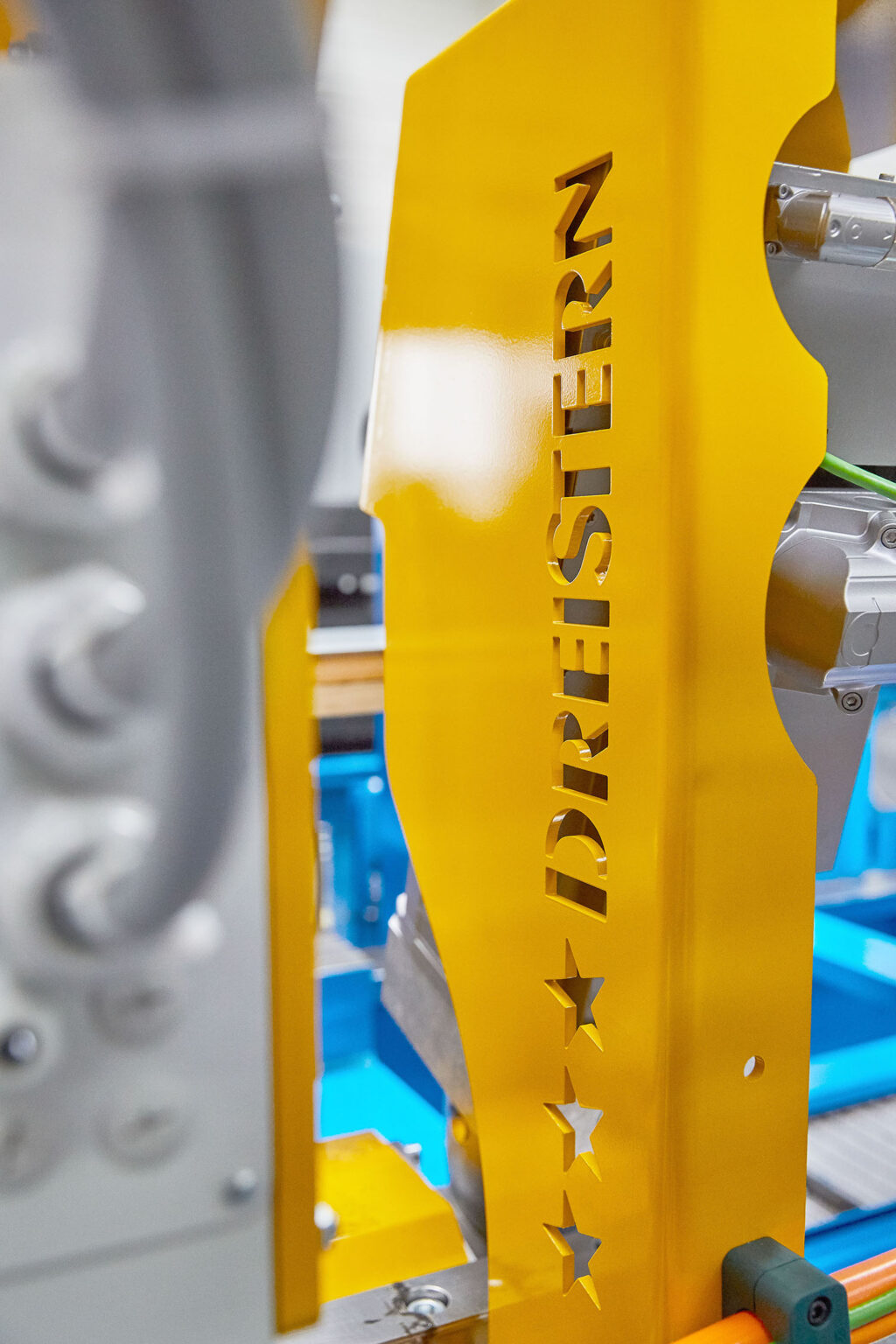 Eine Nahaufnahme eines gelben Metallteils einer industriellen Maschine, auf dem der Schriftzug 'DREISTERN' zusammen mit drei ausgeschnittenen Sternen zu sehen ist. Der Hintergrund zeigt unscharf Teile der Maschine und weitere industrielle Komponenten in Blau und Silber. Das Metallteil ist glänzend und sauber verarbeitet, was auf eine hochwertige Konstruktion hinweist.