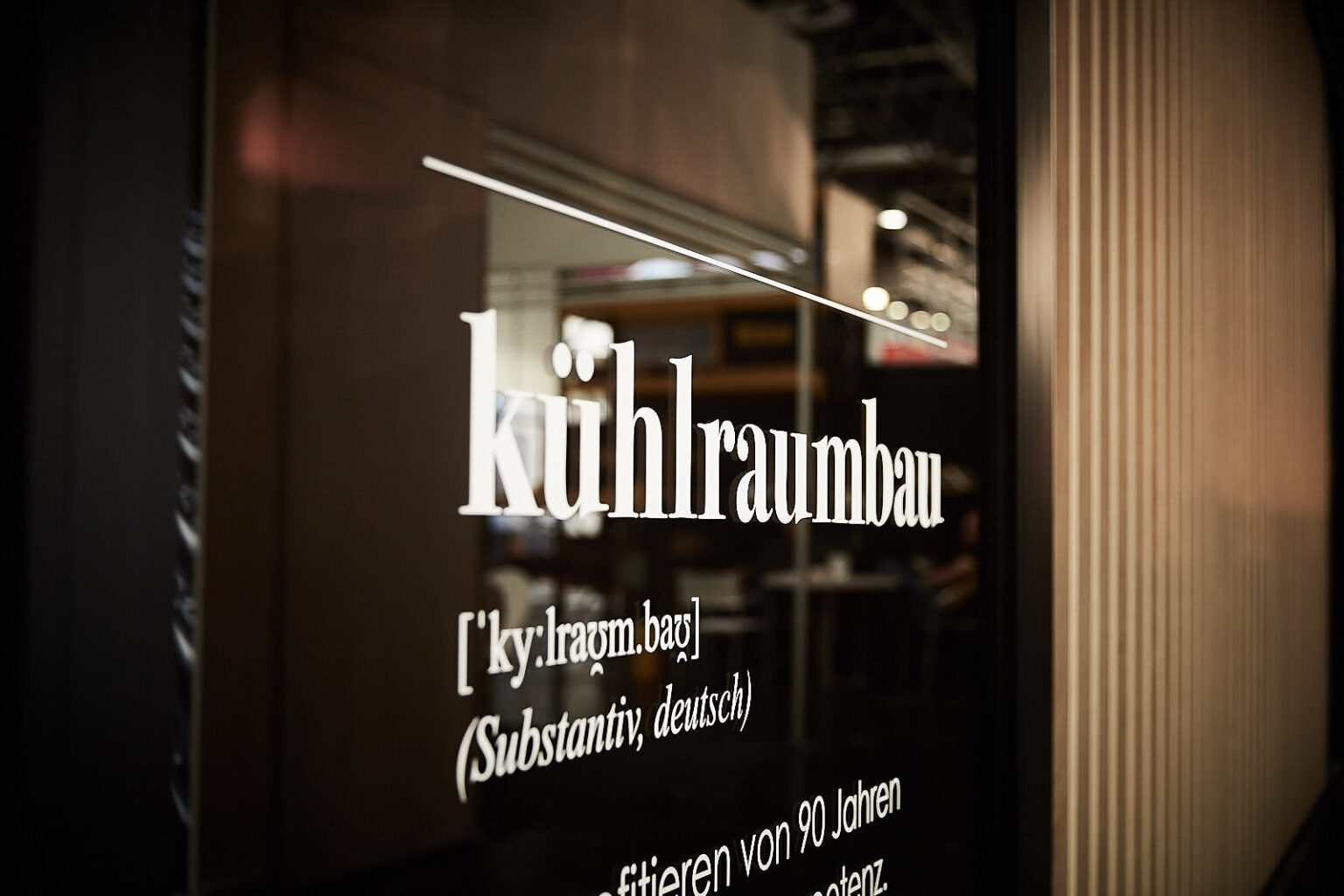 Ein Schild mit der Aufschrift 'kühlraumbau' in weißer Schrift auf dunklem Hintergrund. Unter dem Wort befindet sich die phonetische Aussprache '[ˈky ˌʁaʊmˌbaʊ]' und die Angabe '(Substantiv, deutsch)'. Im Hintergrund ist unscharf eine Messehalle mit weiteren Ständen zu sehen