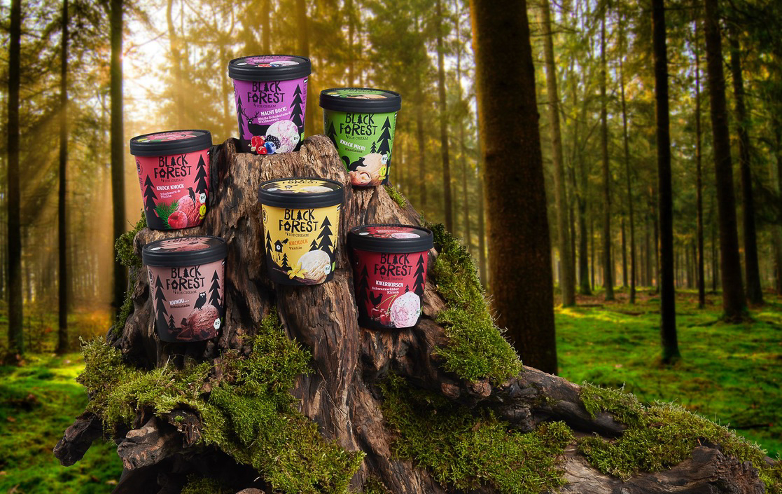 Verschiedene Eissorten der Marke "BLACK FOREST" sind kreativ auf einem alten, moosbewachsenen Baumstumpf in einem Wald drapiert. Die Szene wird von einem weichen, natürlichen Licht beleuchtet, das durch das dichte Blätterdach der hohen Bäume im Hintergrund scheint. Der Boden ist mit leuchtend grünem Moos bedeckt, was einen frischen und lebendigen Eindruck der Natur vermittelt. Die Eissorten sind durch ihre auffälligen Verpackungen mit unterschiedlichen Farben und Fruchtabbildungen gekennzeichnet, darunter Himbeere, Schokolade, Walnuss und Kirsche. Das Arrangement suggeriert einen Genussmoment inmitten der ruhigen und schönen Umgebung des Waldes.