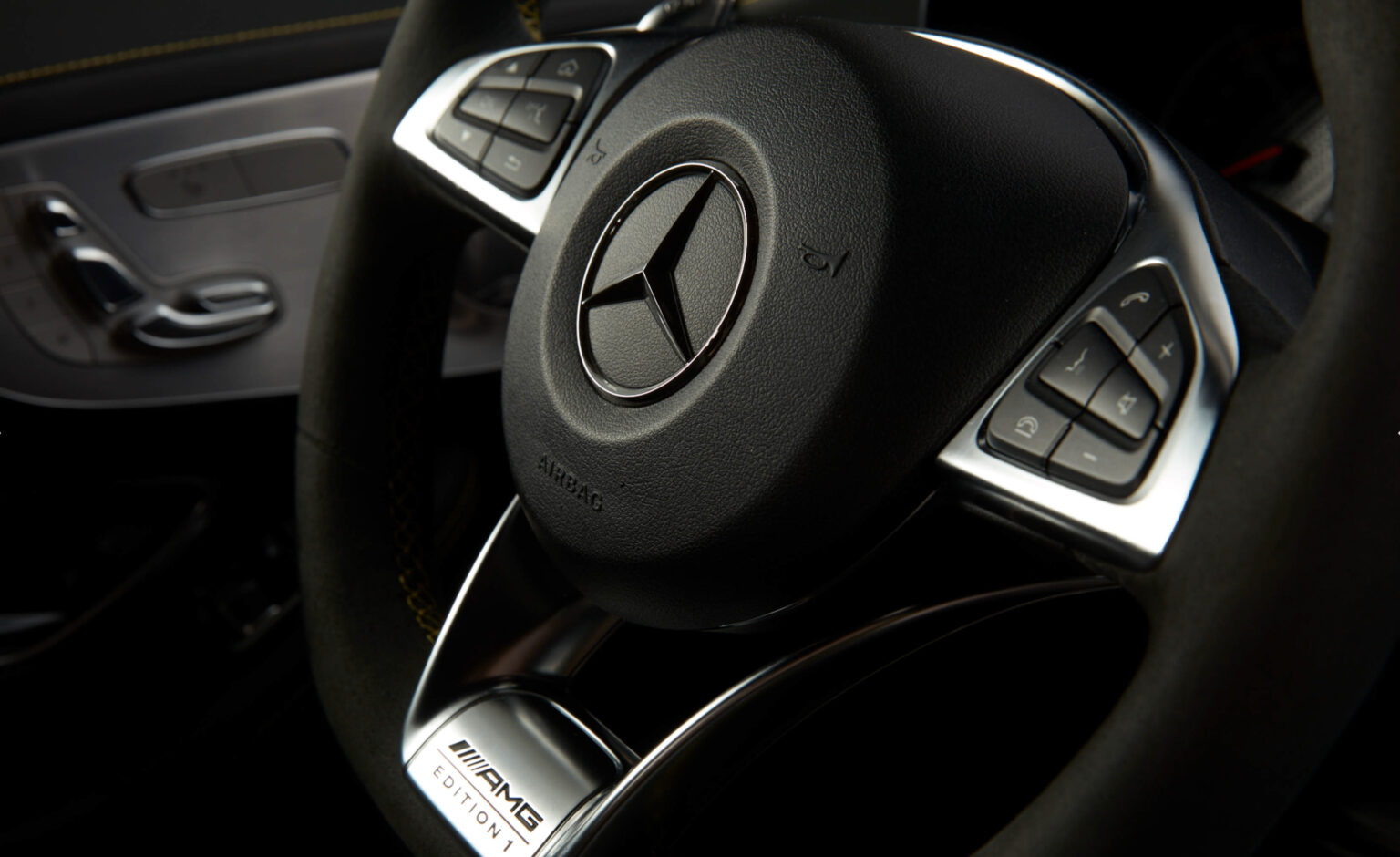 Das Bild zeigt das Lenkrad eines Mercedes-Benz Fahrzeugs, deutlich durch das prominent platzierte Logo in der Mitte gekennzeichnet. Das Lenkrad ist mit dunklem Leder überzogen, und am unteren Rand ist das Logo von AMG mit der Beschriftung "EDITION 1" zu erkennen, was auf eine Sonderedition des Fahrzeugs hinweist. An der Seite des Lenkrads befinden sich silberne Bedienelemente, die wahrscheinlich zur Steuerung von Multimediafunktionen dienen. Das Design ist elegant und vermittelt ein Gefühl von Luxus und sportlicher Ästhetik.