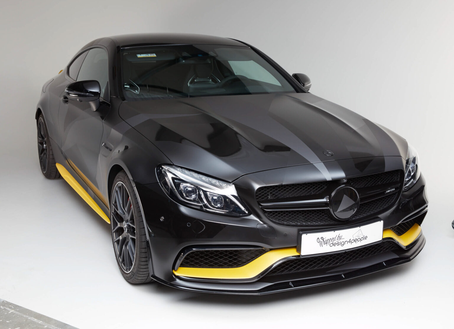 Ein hochglanzschwarzes Mercedes-Benz Coupé steht im Vordergrund, mit markanten gelben Akzentstreifen an der Frontschürze und den Seitenschwellern. Das Auto präsentiert sich in einem Studio mit neutralem Hintergrund. Charakteristisch sind die scharf gezeichneten Scheinwerfer und das glänzende Emblem auf dem Kühlergrill, die die hochwertige Verarbeitung und das sportliche Design des Fahrzeugs betonen. Auf dem Nummernschild steht "Wrapped by Design4People", was darauf hinweist, dass das Auto eine individuelle Folierung erhalten hat.