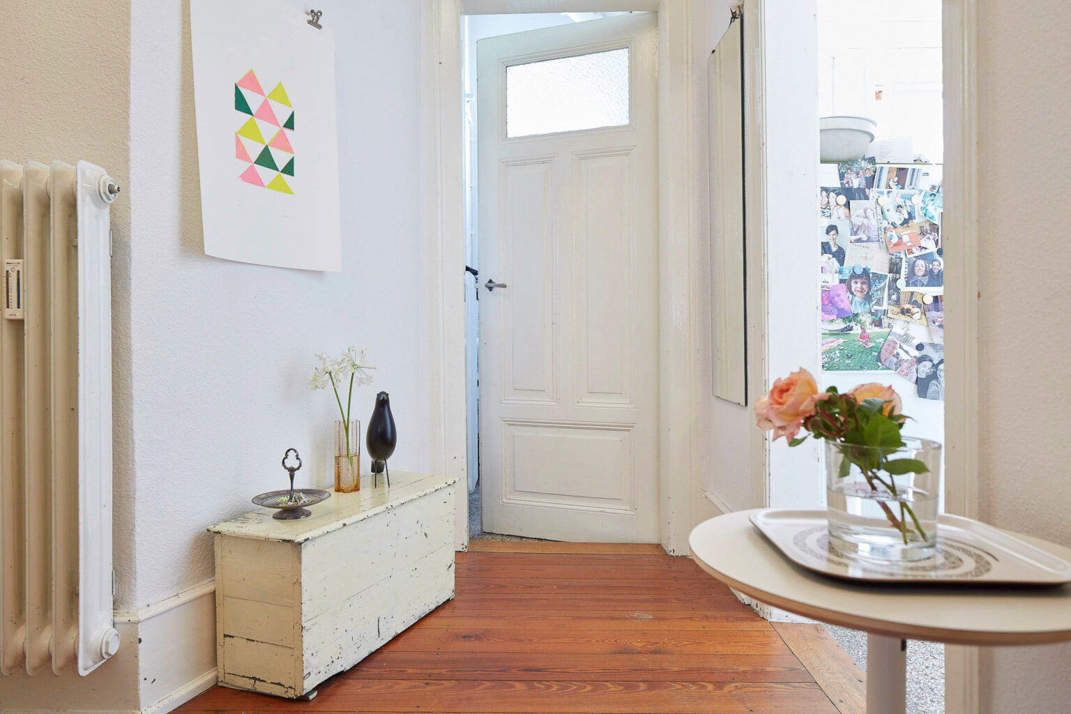 Ein heller Flur mit weiß gestrichenen Wänden und Türen. Der Boden ist mit Holzplanken ausgelegt. Auf der linken Seite steht eine kleine, weiß gestrichene Truhe, auf der eine Vase mit weißen Blumen und eine dekorative Schale steht. Daneben befindet sich ein schwarz lackierter Vogel als Deko. An der Wand darüber hängt ein Bild mit bunten Dreiecken. Rechts im Bild ist ein kleiner Tisch mit einer Vase mit rosafarbenen Blumen zu sehen. Im Hintergrund ist ein Raum mit vielen Fotos an einer Pinnwand sichtbar.