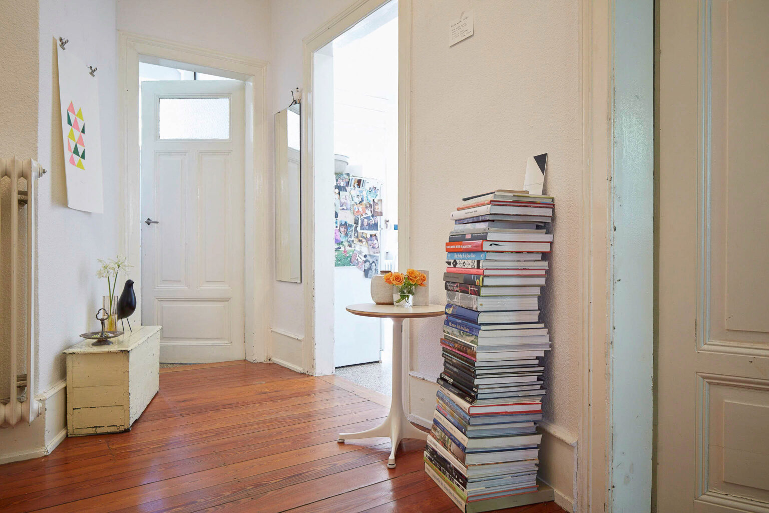 Ein heller Flur in einer Wohnung mit Holzboden. An der linken Wand hängt ein farbenfrohes Bild und ein Spiegel, daneben steht eine weiße Truhe mit einer Vase und einer schwarzen Dekofigur. In der Mitte des Flurs steht ein kleiner runder Tisch mit einem Blumenstrauß. Rechts ist ein hoher Stapel Bücher, der bis auf Tischhöhe reicht. Im Hintergrund ist eine Tür zum nächsten Raum zu sehen, in dem ein Kühlschrank mit vielen Fotos und Notizen an der Tür steht.