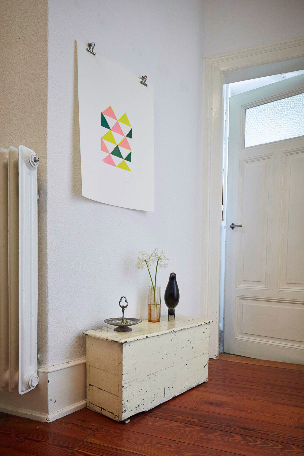 Ein heller Flur mit weiß gestrichenen Wänden und einer geschlossenen, weißen Tür im Hintergrund. Auf dem Holzboden steht eine alte, weiß gestrichene Truhe mit abgeblätterter Farbe. Darauf befinden sich eine Vase mit weißen Blumen, eine kleine Metallplatte und eine schwarze Vogelskulptur. An der Wand über der Truhe hängt ein Bild mit bunten Dreiecken. Links im Bild ist ein weißer Heizkörper zu sehen