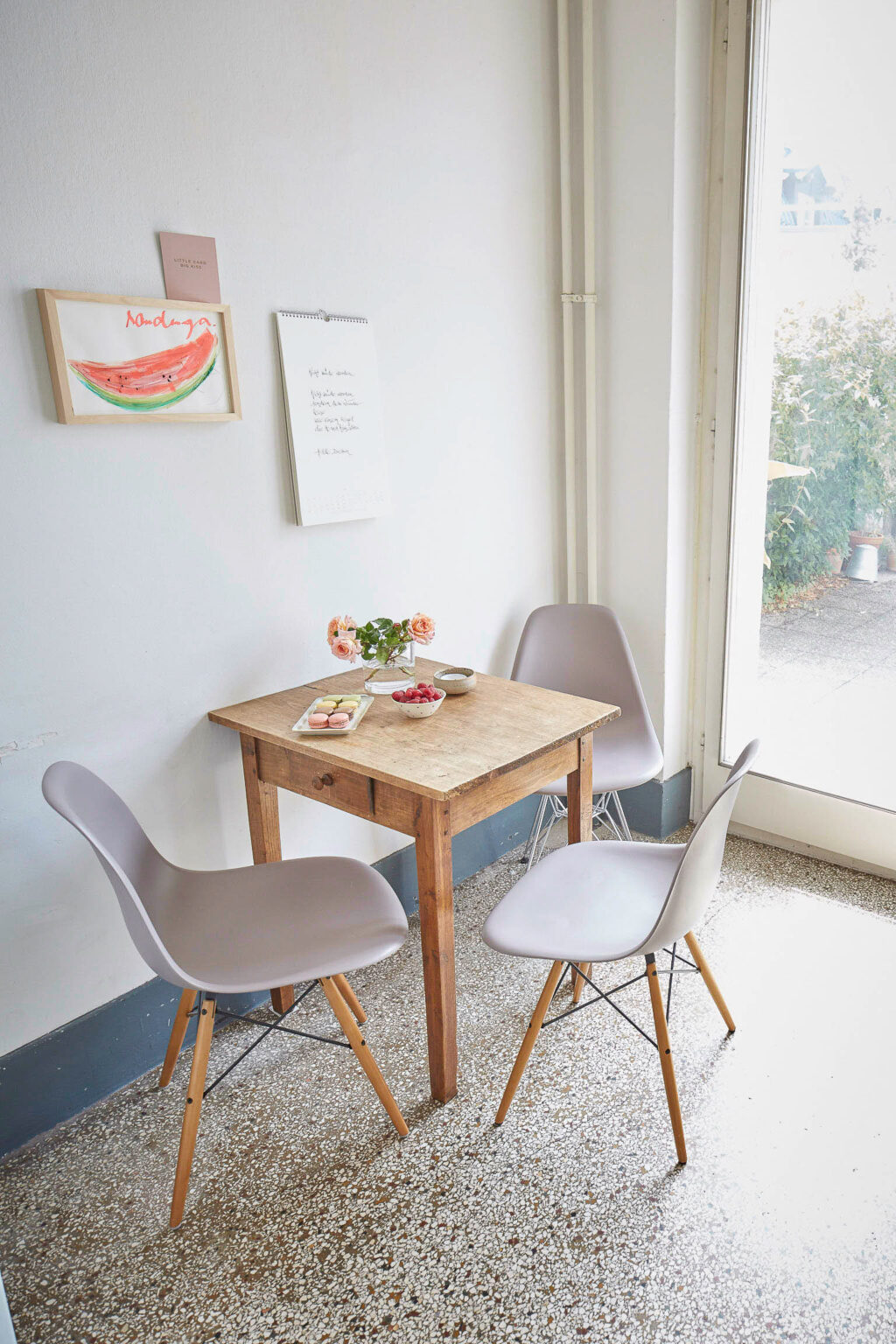 Ein kleiner, rustikaler Holztisch mit drei modernen grauen Stühlen steht in einer hellen Ecke eines Raumes. Auf dem Tisch befinden sich eine Vase mit Rosen, eine Schale mit Himbeeren und ein Teller mit Macarons. An der Wand hängen ein Bild eines Wassermelonenstücks und ein Kalenderblatt mit handgeschriebenem Text. Durch eine Glastür rechts im Bild fällt Tageslicht in den Raum, und draußen sind Pflanzen zu sehen.